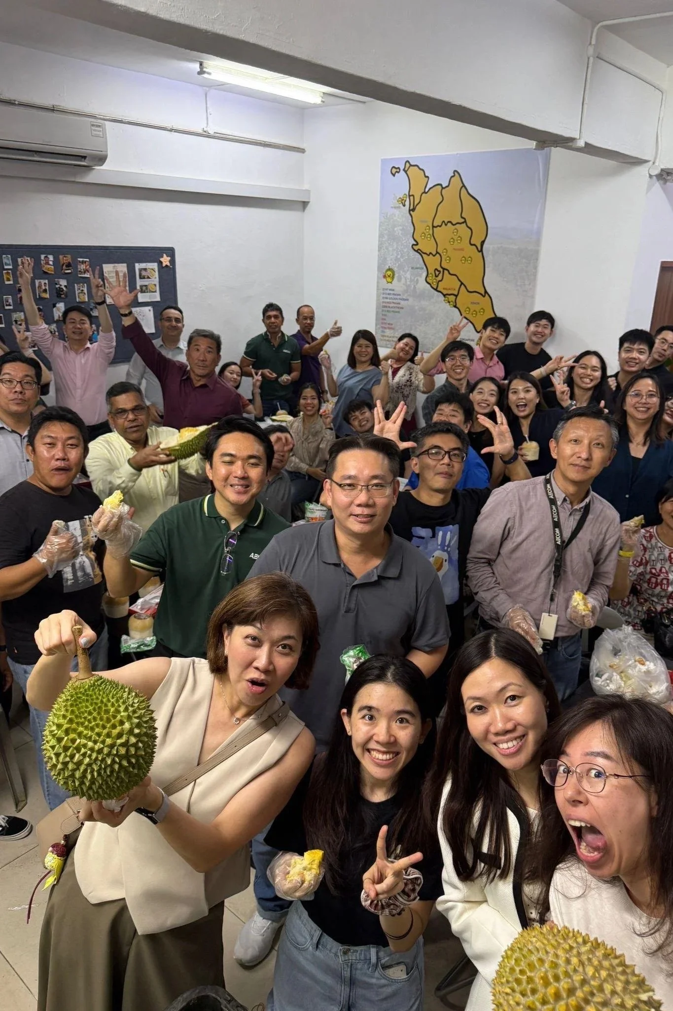 A large group of people gathered in a room, smiling and making peace signs, with some holding durian fruit. There is a map of Thailand on the wall behind them, and a bulletin board with photos and postcards. The group appears to be at a social or cultural event.