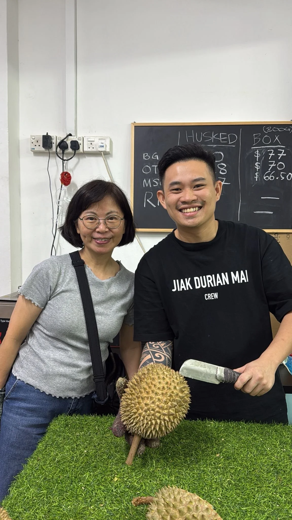Two people smiling next to durian fruit on a green table, with a blackboard in the background.