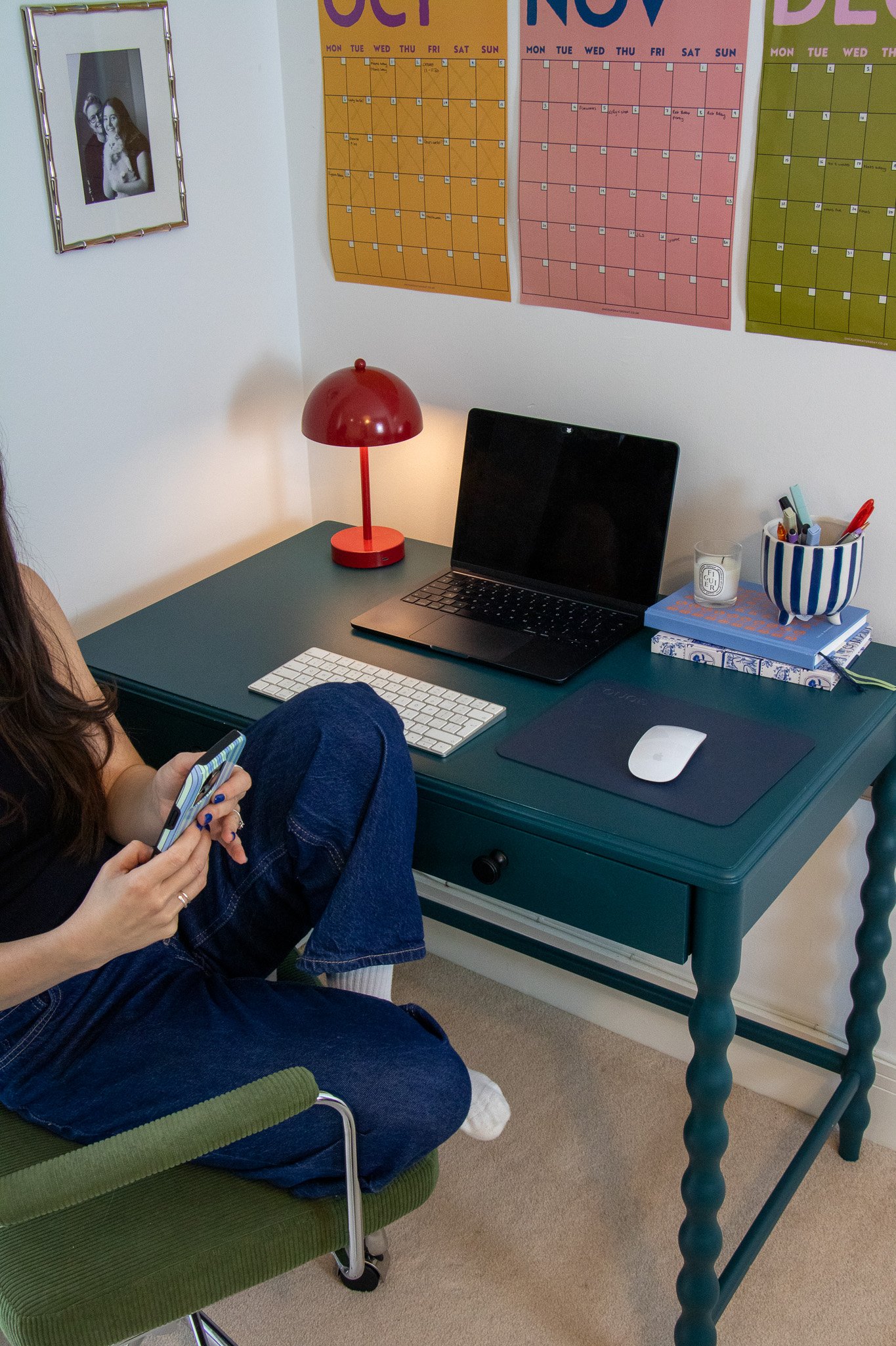 A person sitting on a green chair at a desk with a laptop, keyboard, mouse, and a red desk lamp, in a room with colorful wall calendars and a framed black-and-white photo.