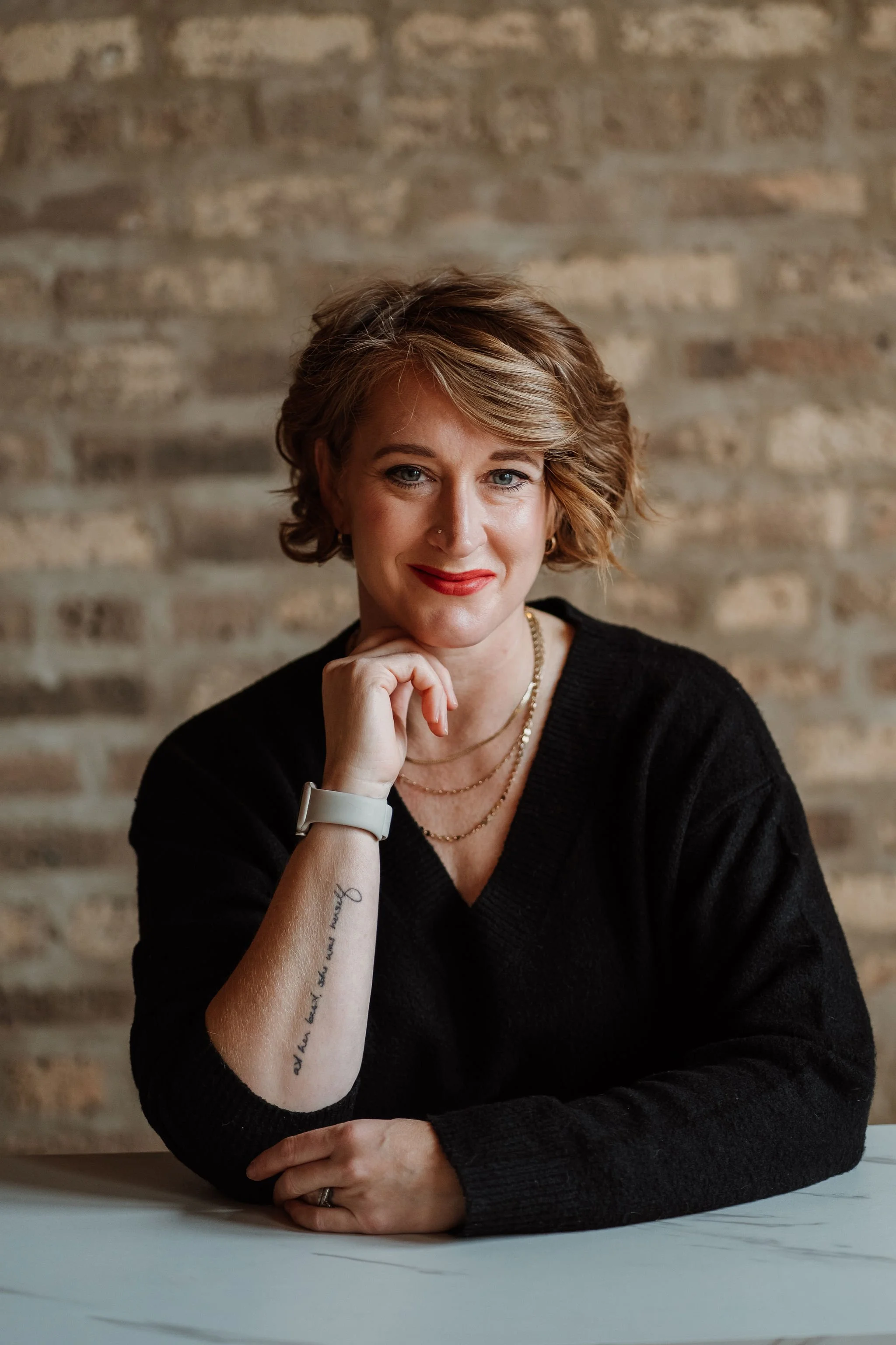 A woman with short, wavy red hair and blue eyes seated at a desk against a brick wall background. She has a small nose piercing, wears red lipstick, and is dressed in a black sweater. She rests her chin on her hand, with her elbow on the desk, wearing a white watch on her left wrist and layered gold necklaces. She has a tattoo on her right forearm.