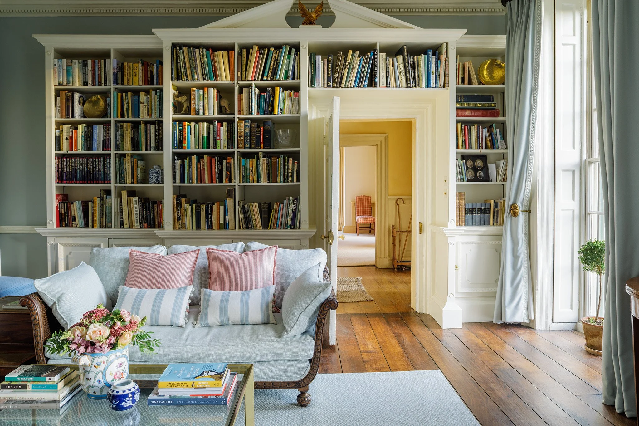 A cozy living room with a white sofa decorated with pastel pillows, a glass coffee table with books and a flower vase, large open bookcases filled with books and decorative items, and large windows with white curtains allowing natural light.