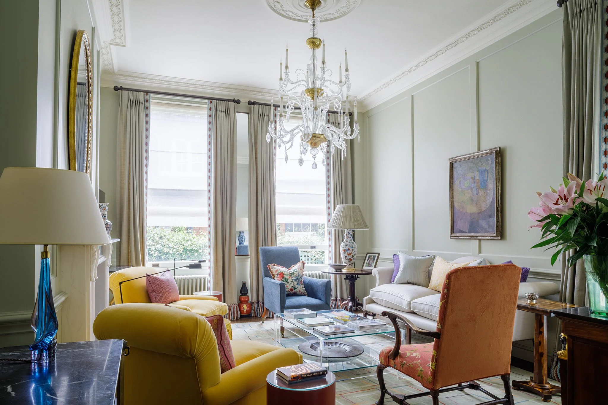 Living room with green paneled walls, featuring a white sofa, a blue armchair, an orange armchair, a yellow armchair with pink cushions, a glass coffee table with books, a chandelier hanging from the ceiling, large windows with cream curtains, and de