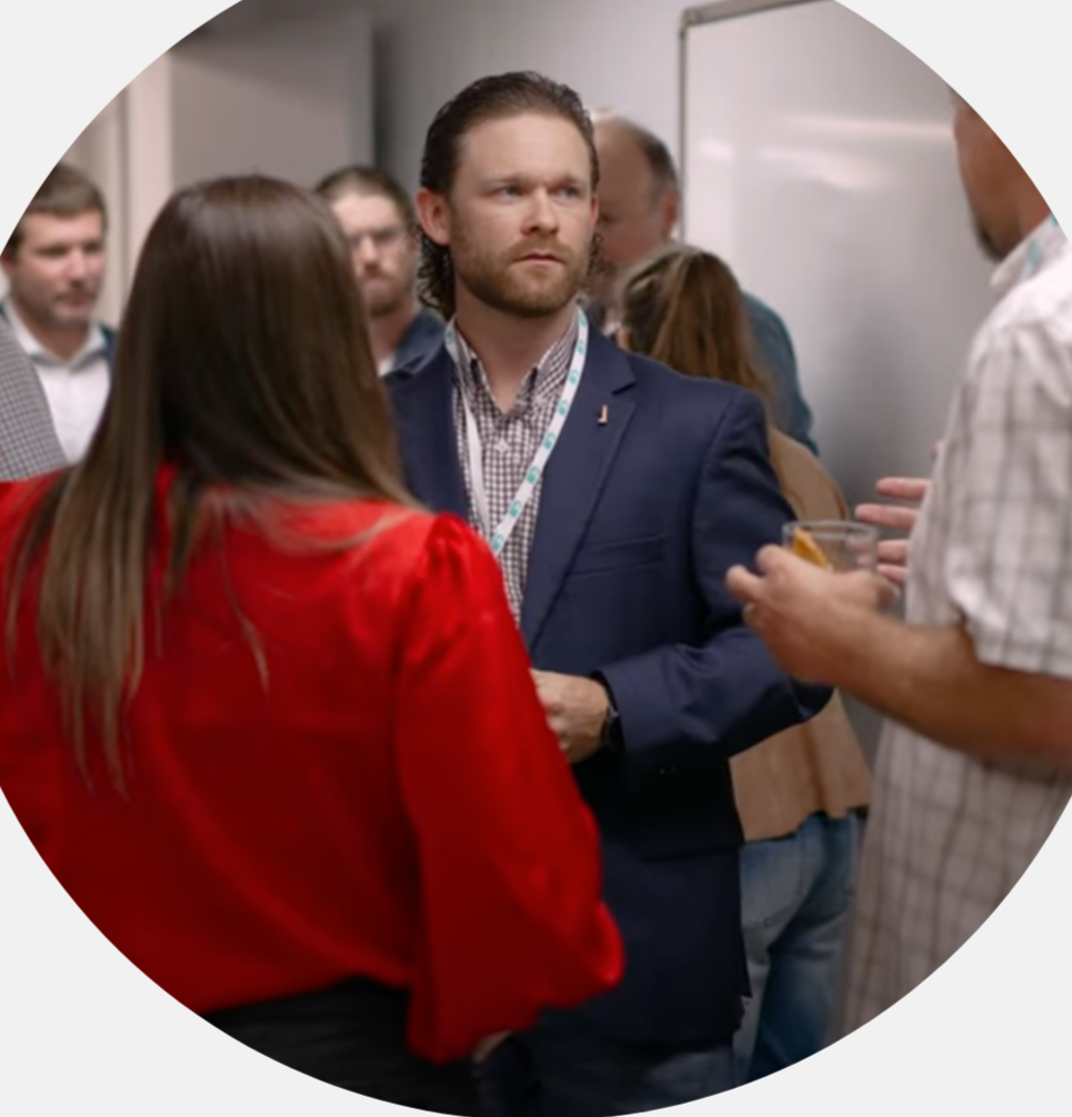 People engaged in conversation at a social event, with a focus on a man in a navy blazer and patterned shirt listening to a man in a checkered shirt holding a drink in front of a whiteboard.