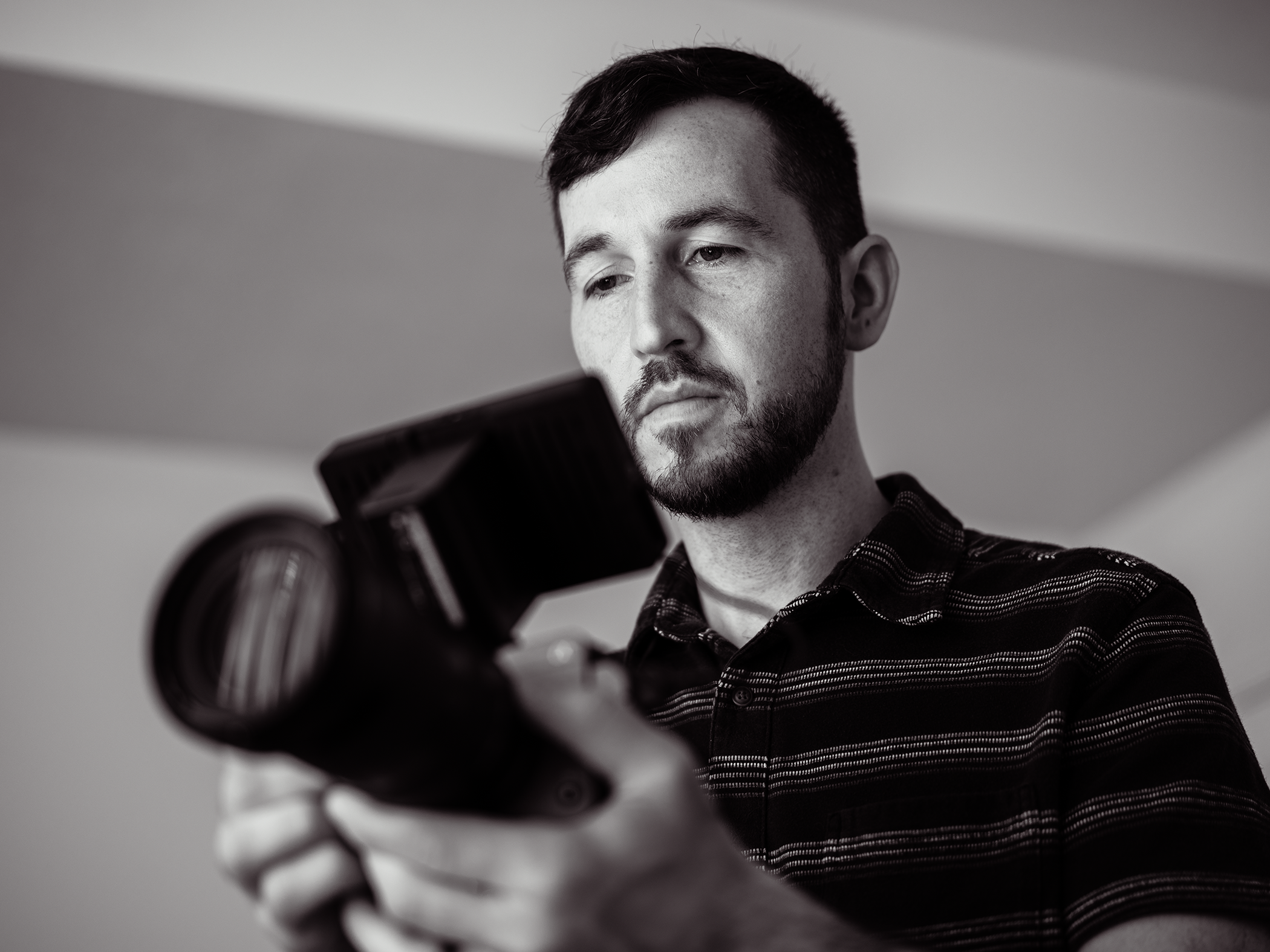 A man with dark hair and a beard, wearing a striped shirt, looks at a camera he is holding.