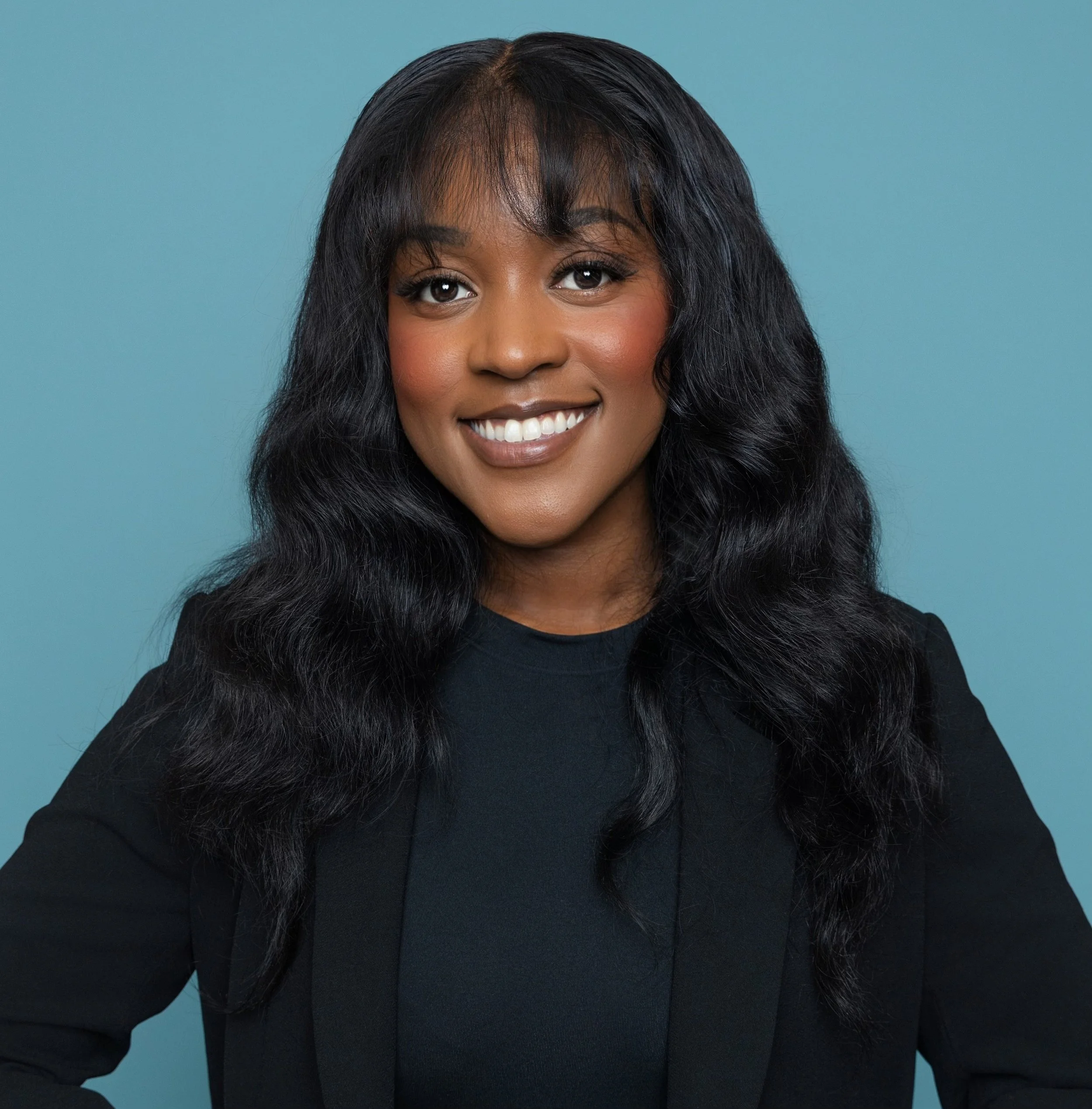 A woman with long black wavy hair smiling against a blue background.