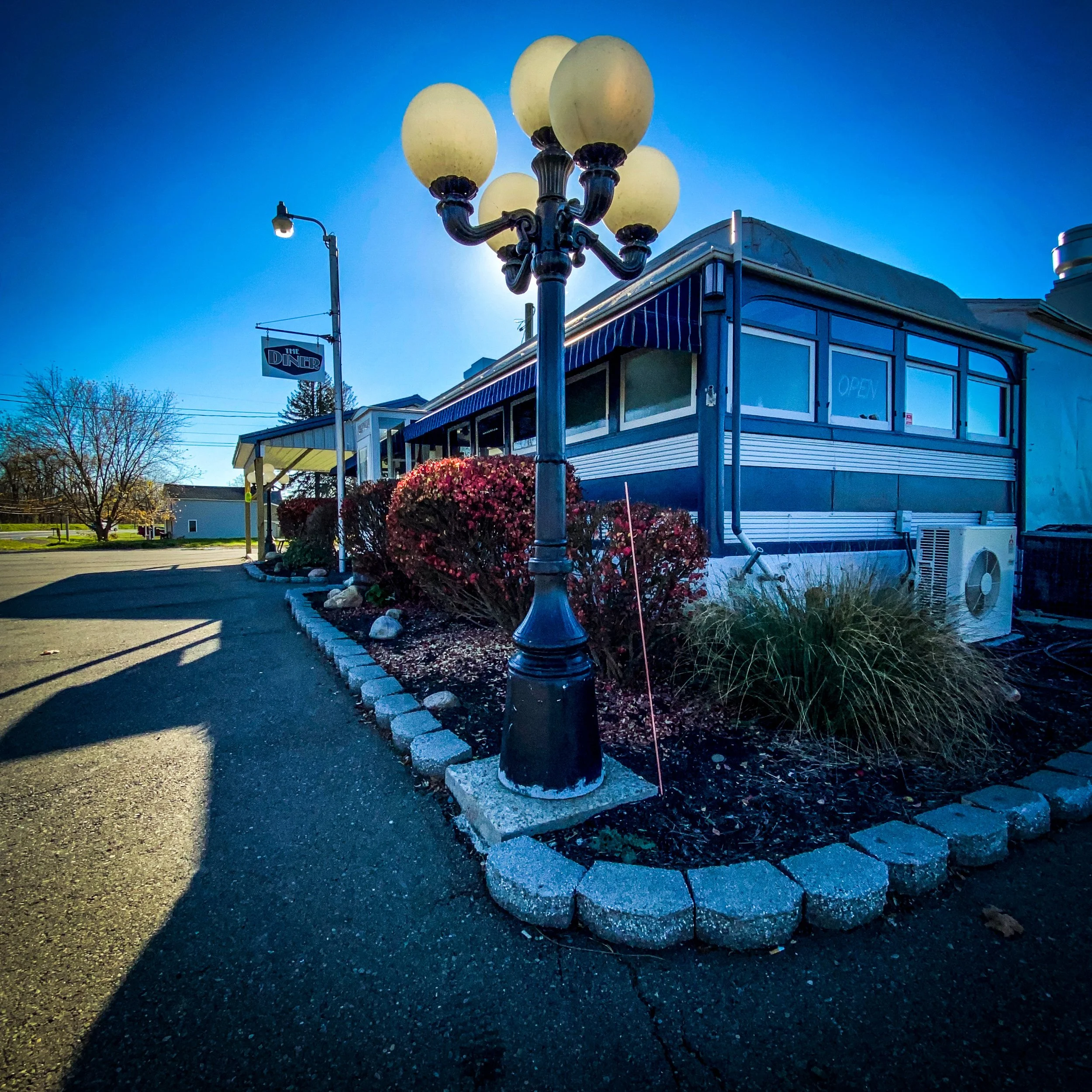 Exterior view of a vintage diner with a striped awning and glass windows, featuring a black lamppost with spherical lights in the foreground, surrounded by landscaping with bushes and grass.