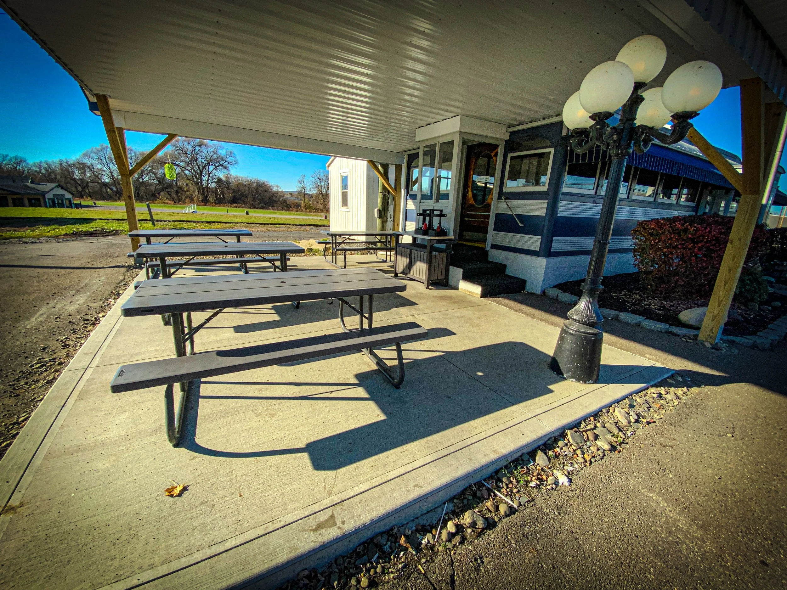 Outdoor seating area with picnic tables under a metal canopy, next to a building with a vintage design and a multi-globe lamp post.