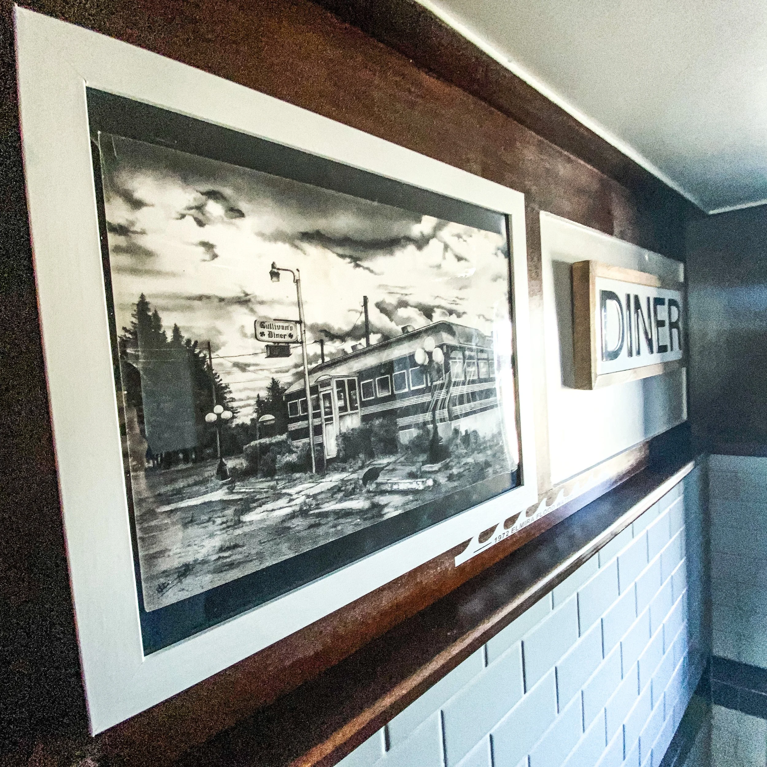 Framed artwork depicting a vintage diner with a cloudy sky, hung on a wall next to a wooden "DINER" sign in a hallway.