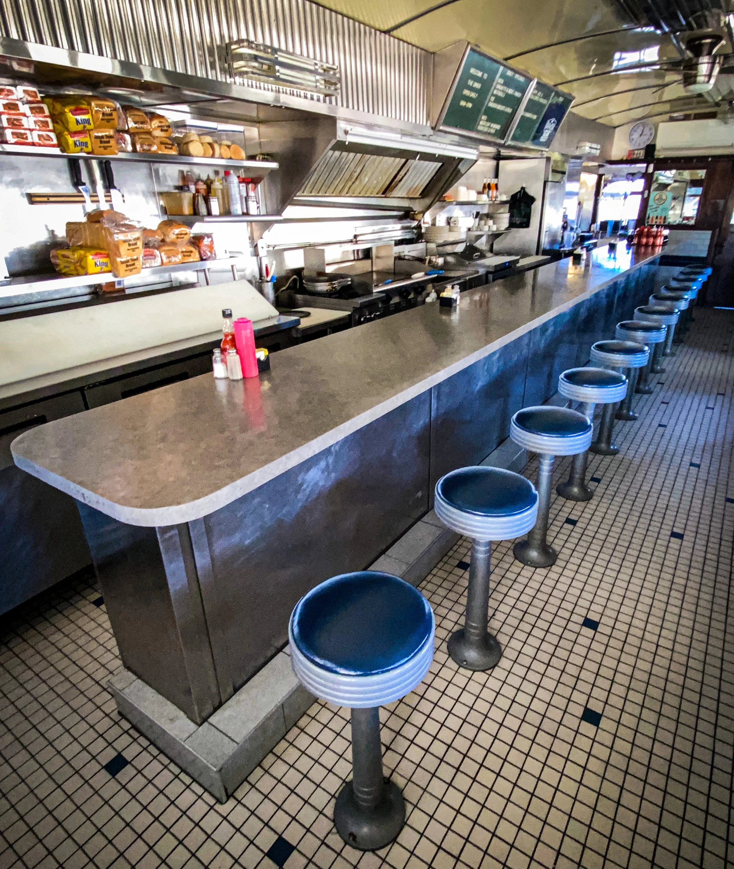 Interior of a classic American diner with a long counter, retro stools, and assorted condiments. Shelves hold bread and burger buns; a cooking area is in the background.