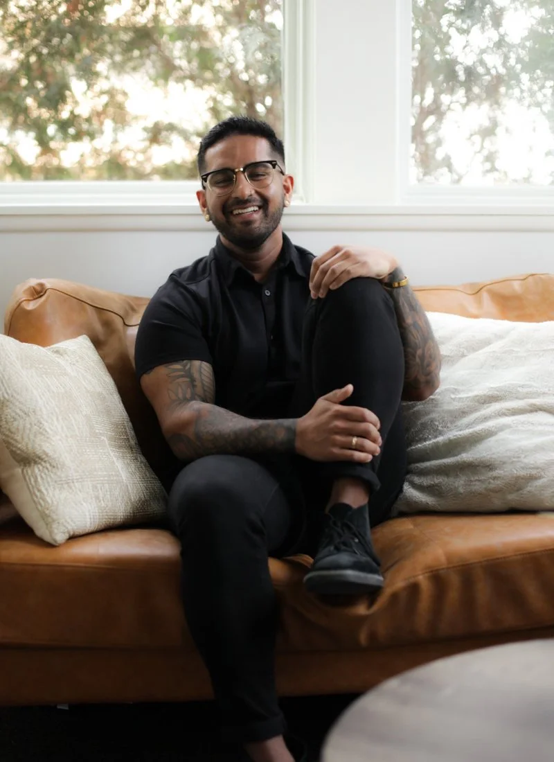 A man with glasses, tattoos, and a dark shirt sitting on a tan leather sofa with cushions, smiling at the camera in a well-lit room with large windows showing trees outside.