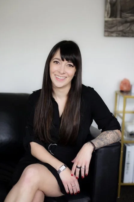 A woman with long dark hair and bangs sitting on a black couch, smiling, wearing a black dress with rolled-up sleeves, a watch, and a ring, in a room with white walls and a yellow side table.