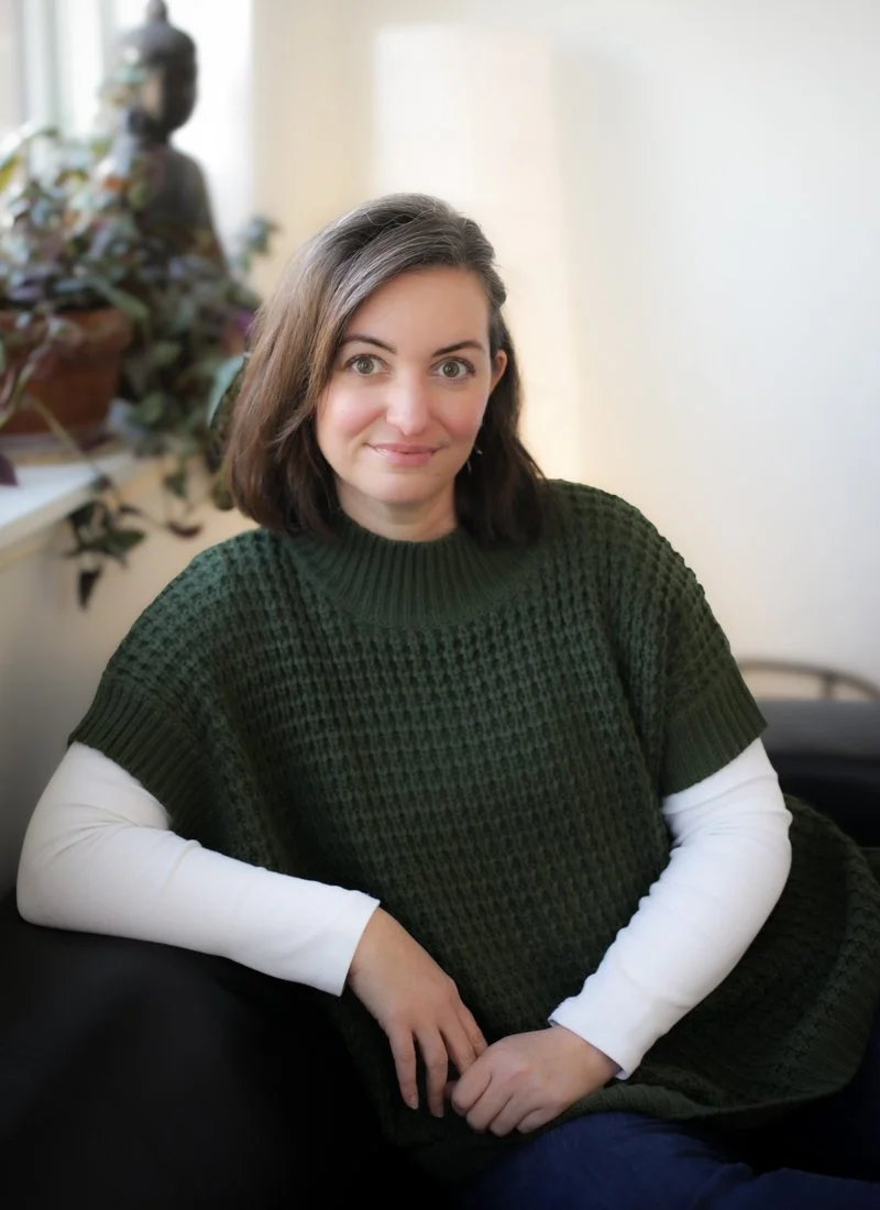 A woman with shoulder-length brown hair, wearing a dark green sweater over a white long sleeve shirt, smiling while sitting on a black chair in a room with a potted plant and a window in the background.