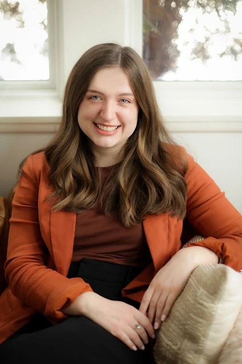A young woman with long, wavy brown hair, smiling, sitting on a beige sofa in front of a window with white frames.