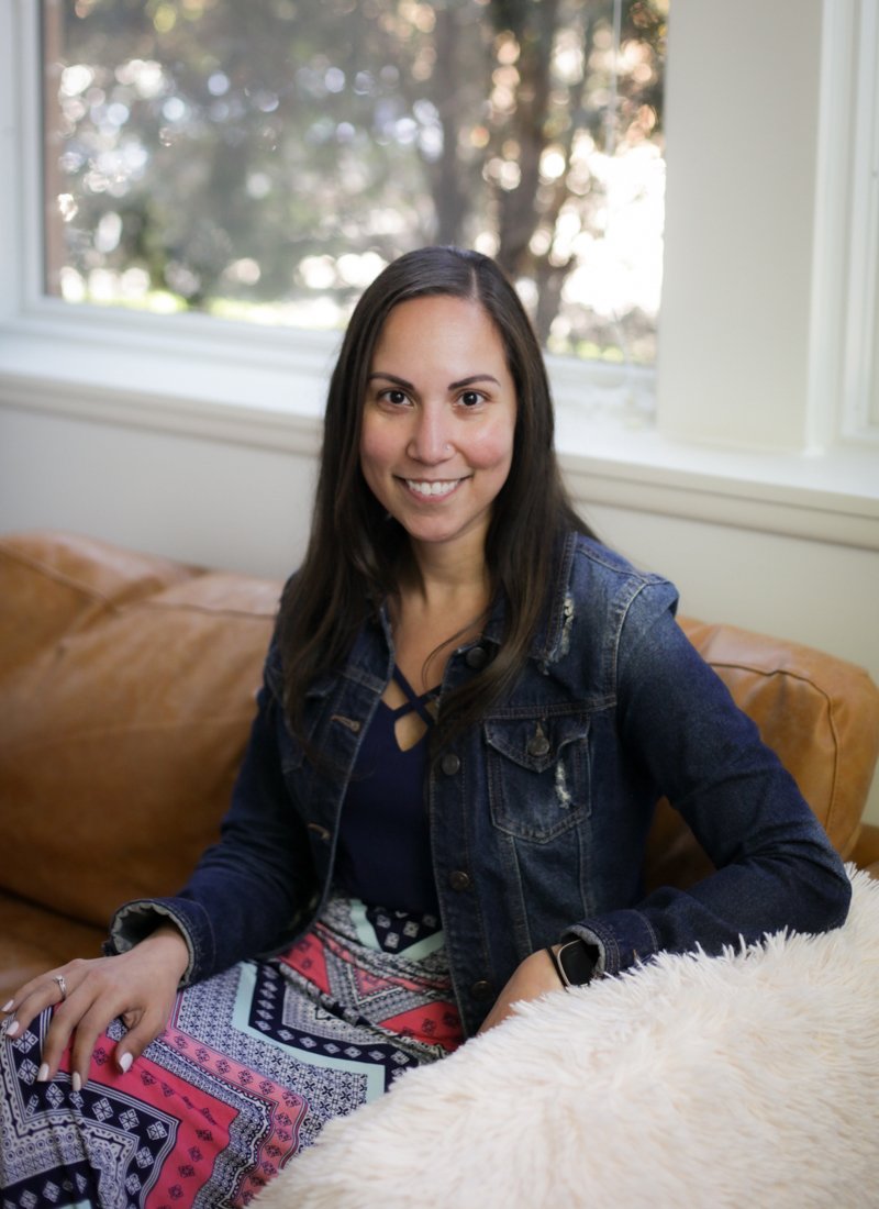 A woman sitting on a brown leather couch near a window, smiling at the camera, wearing a denim jacket and patterned skirt.
