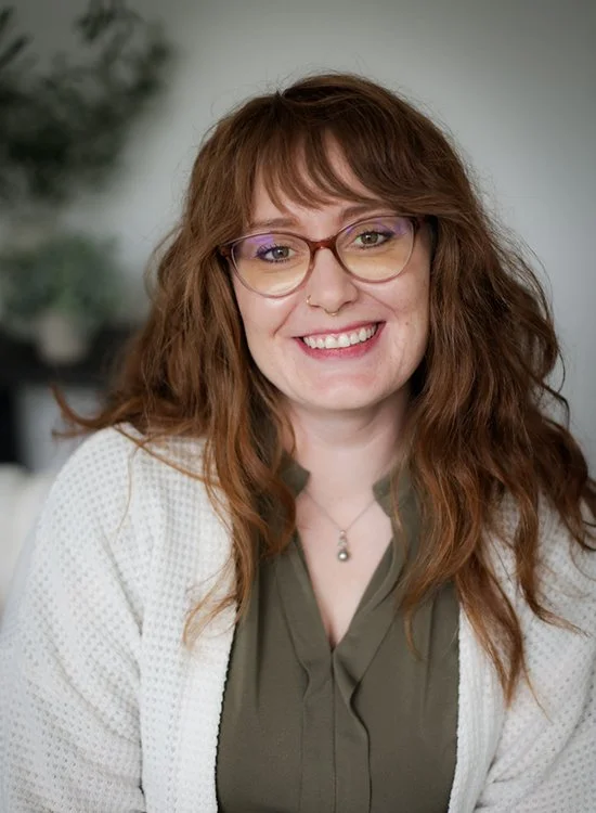 A young woman with wavy red hair, glasses, and a nose piercing, smiling at the camera. She is wearing a green blouse and white cardigan, with a blurred indoor background.
