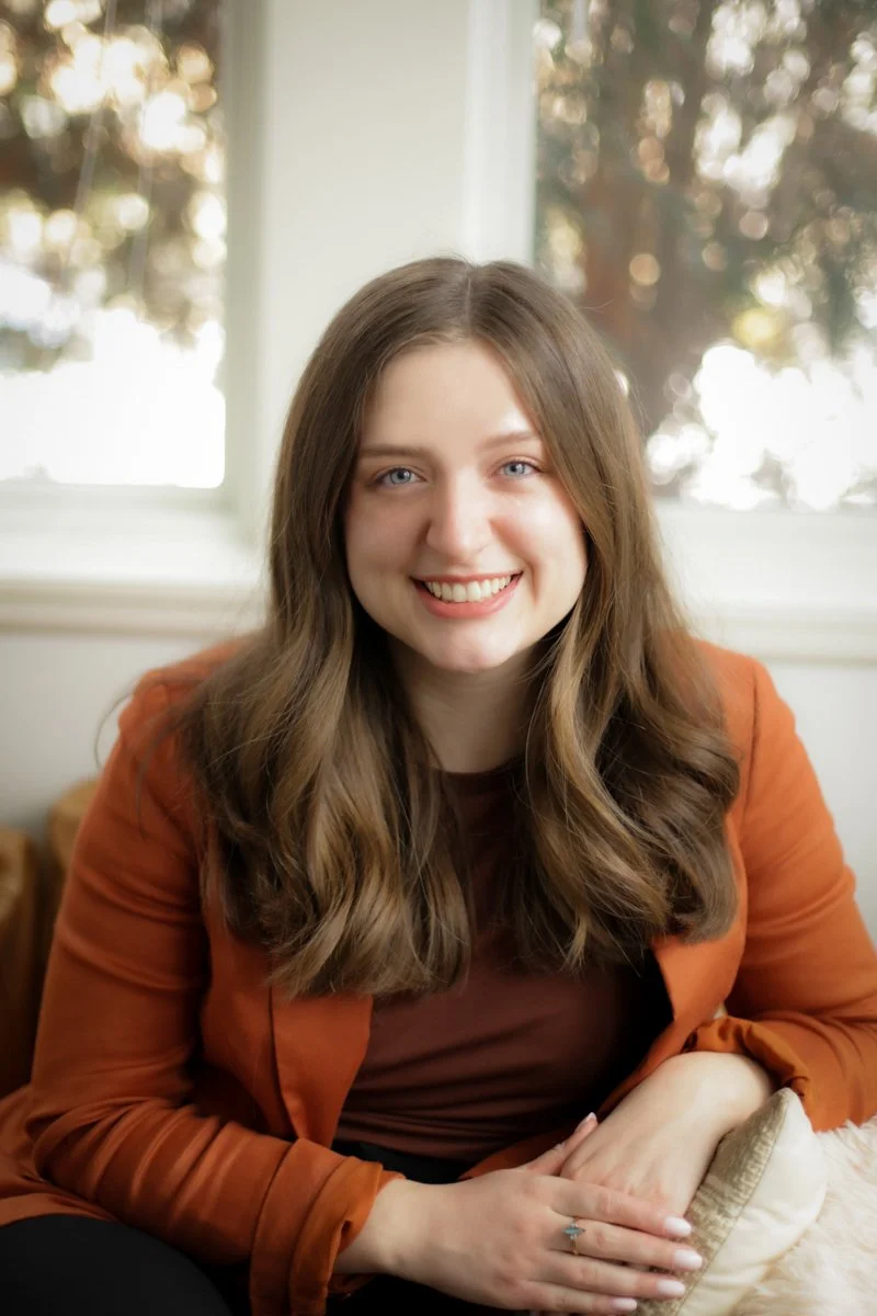 A woman with long wavy brown hair, smiling, seated indoors near a window with trees outside.