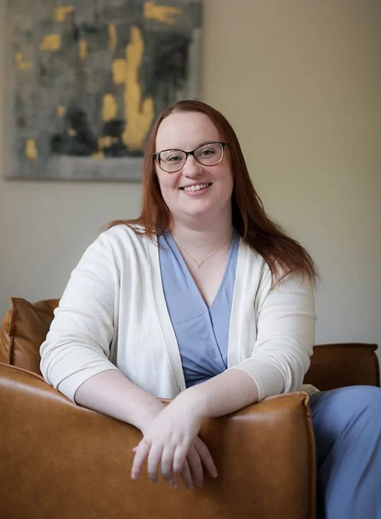 A woman with red hair and glasses, wearing blue scrubs and a white cardigan, sitting on a leather couch with a smile in a room with beige walls and a decorative abstract painting.