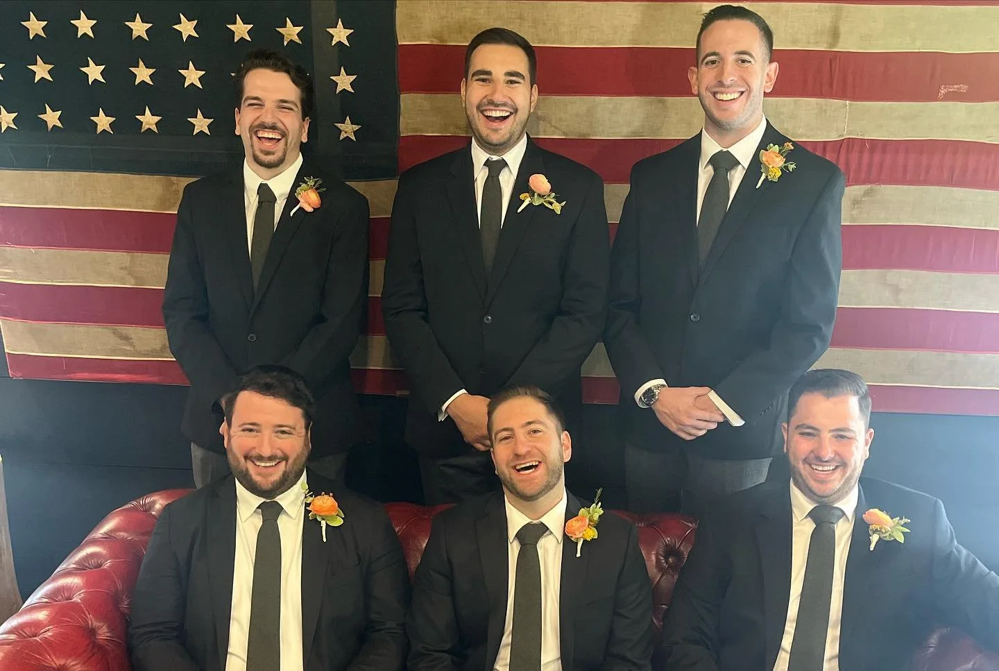 Six men in suits with boutonnieres posing in front of an American flag, some sitting on a red leather couch.