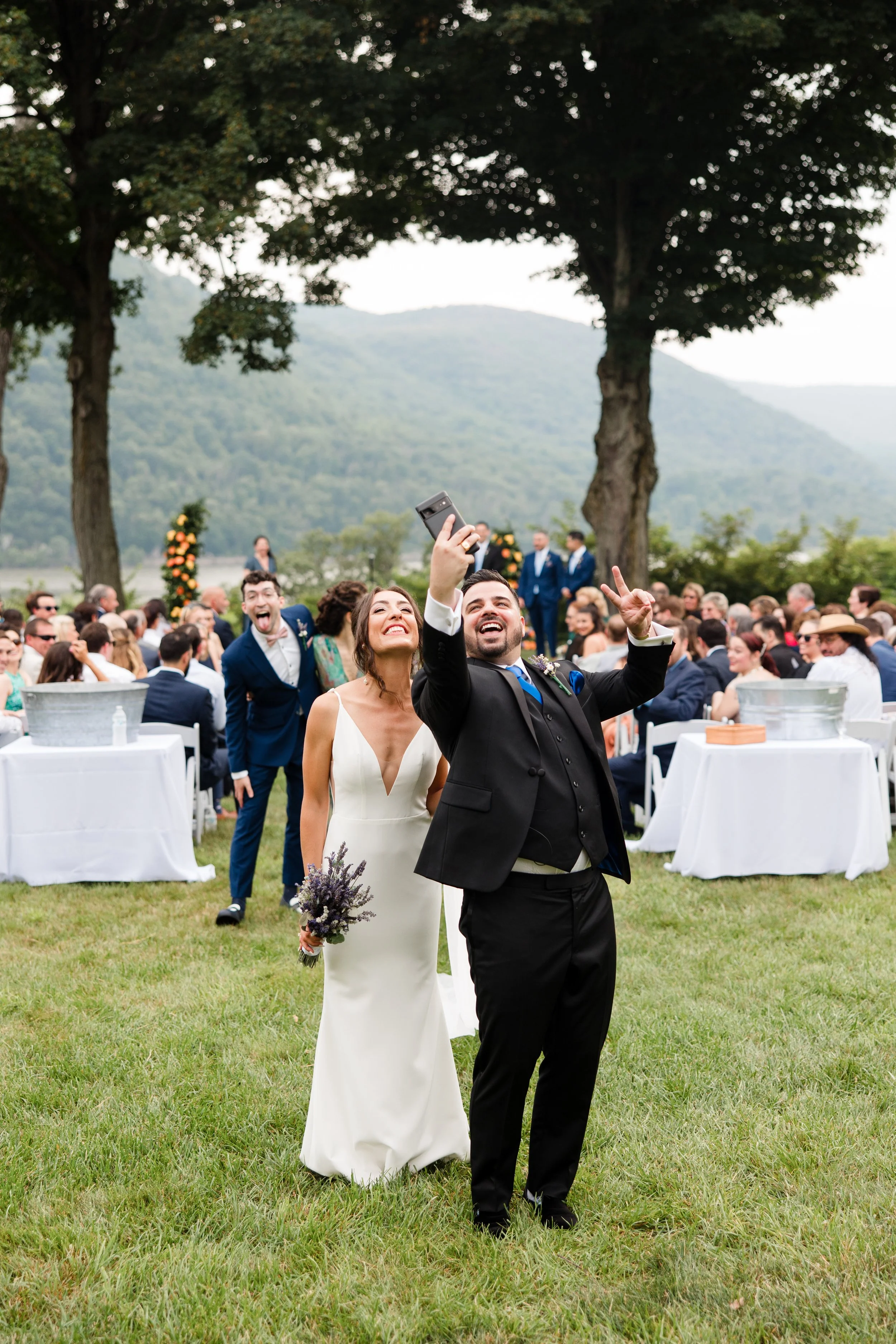 A wedding scene in a grassy outdoor setting with a bride and groom taking a selfie. The bride is wearing a white gown and holding flowers, while the groom is in a black suit. A man in a blue suit makes a playful gesture in the background. Guests are 