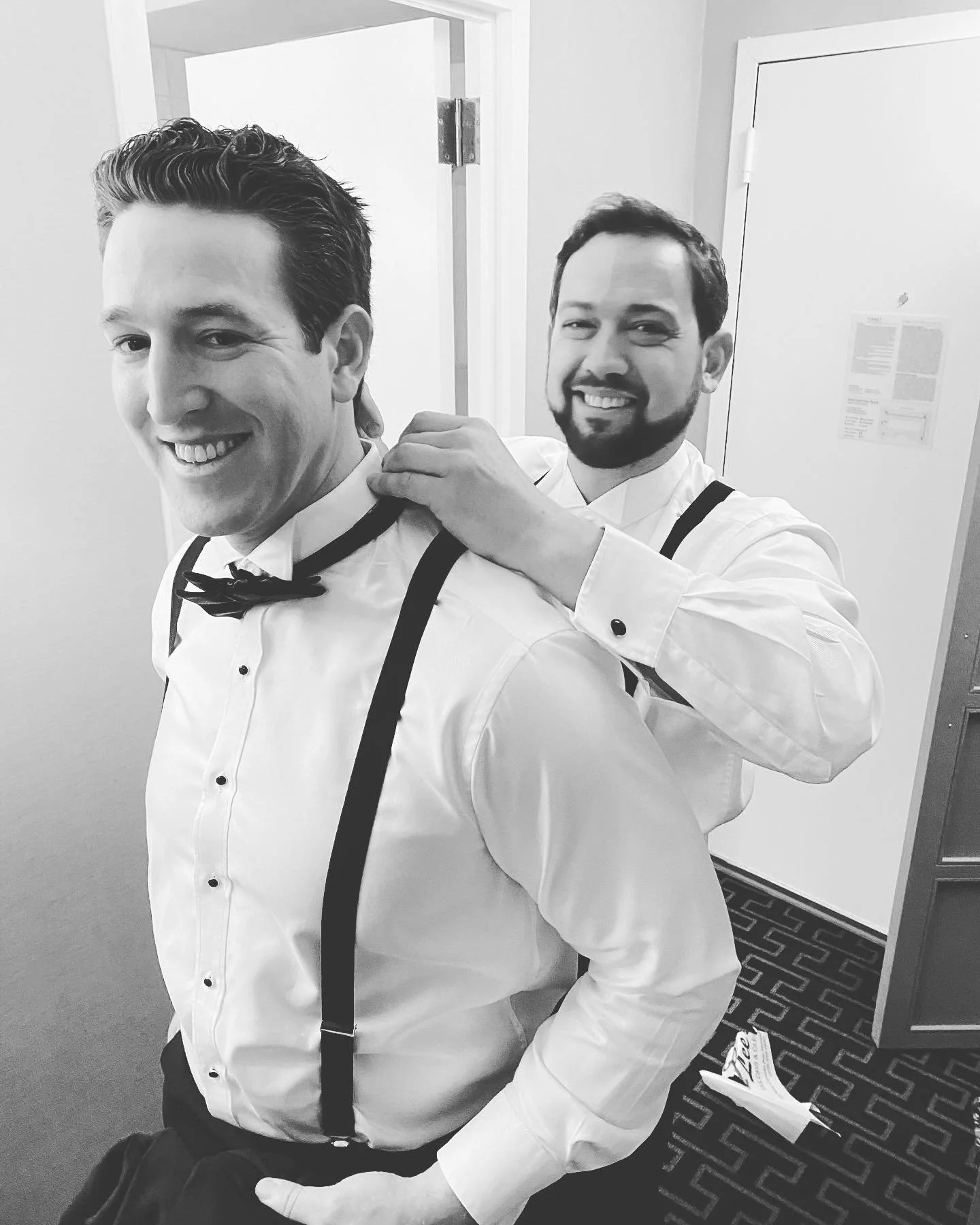 Two men in formal attire, one adjusting the other's bow tie, black and white photo.