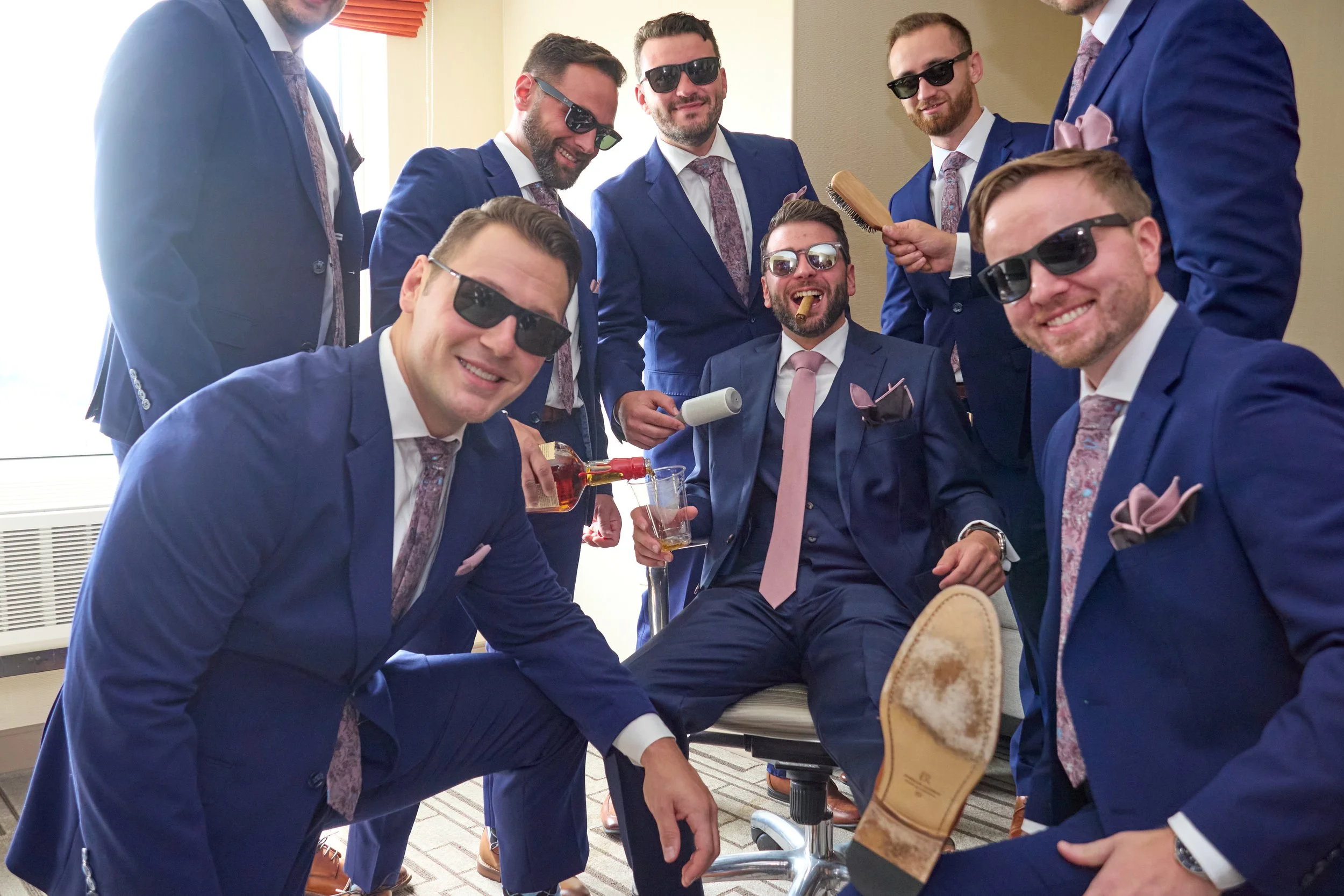 Group of men in blue suits with pink ties and pocket squares, wearing sunglasses, laughing and having fun in a room.