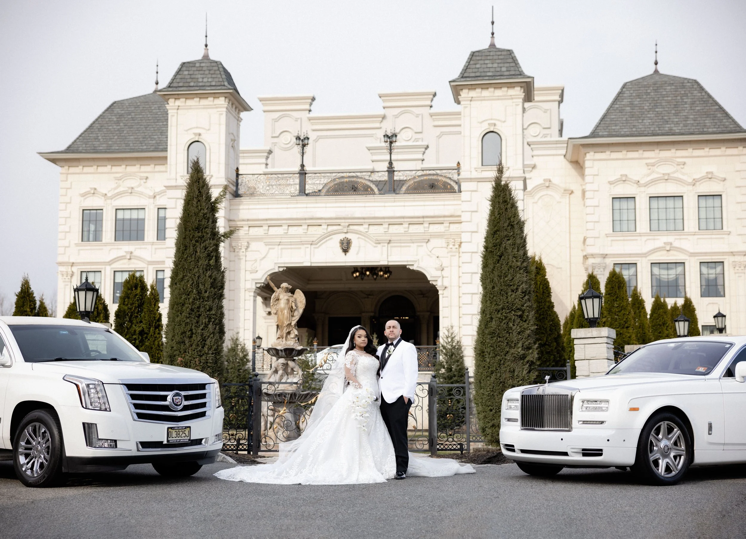 A bride and groom standing outside a grand building with architectural details. They are dressed in formal wedding attire, with two luxury cars, a Cadillac and a Rolls-Royce, parked on either side. The entrance is flanked by tall, conical trees and a