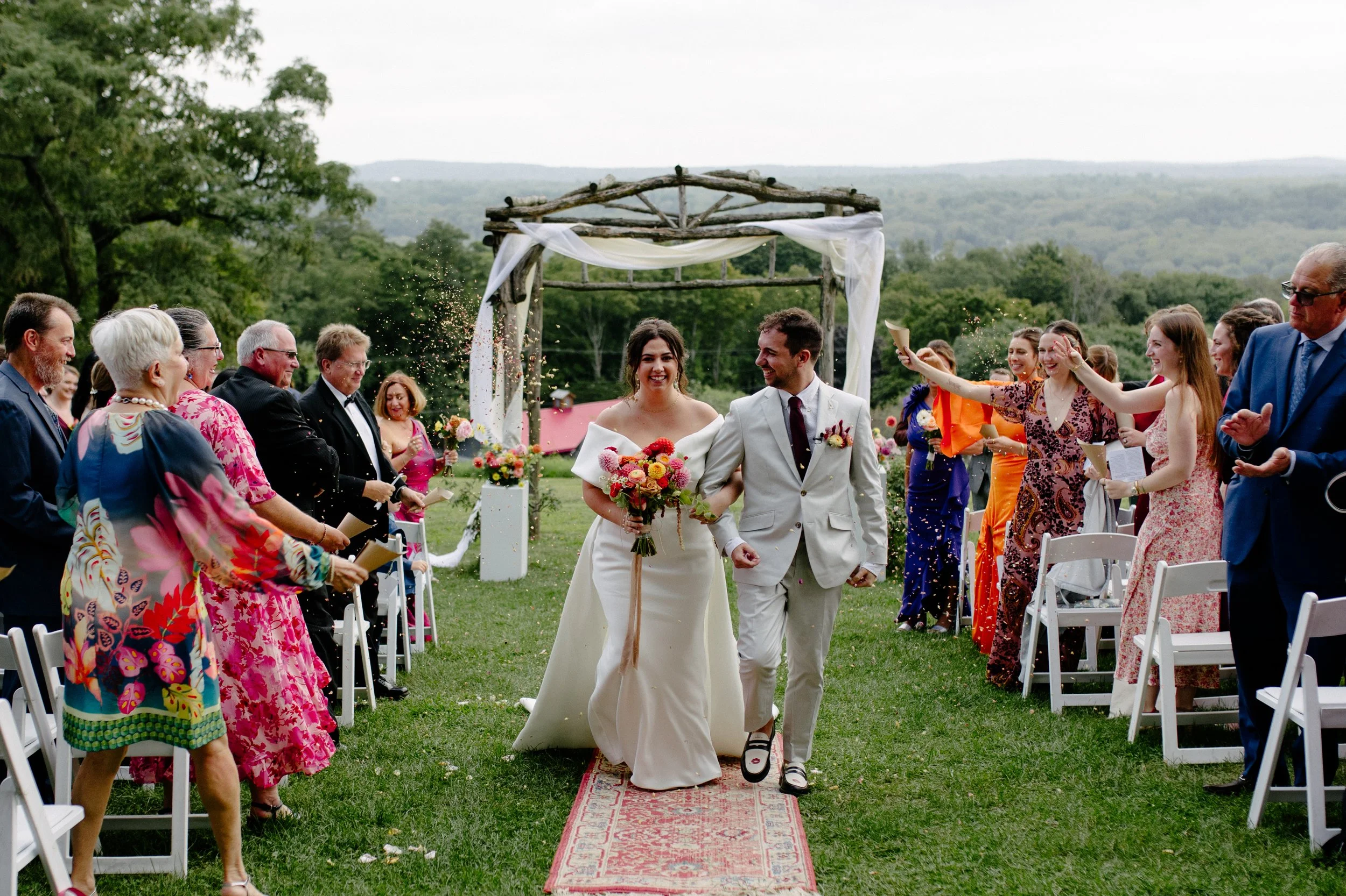 A bride and groom walking down an aisle outdoors after their wedding ceremony, with guests standing and clapping. The bride is wearing a white wedding gown, holding a bouquet, and the groom is in a light suit. There's a wooden archway decorated with 