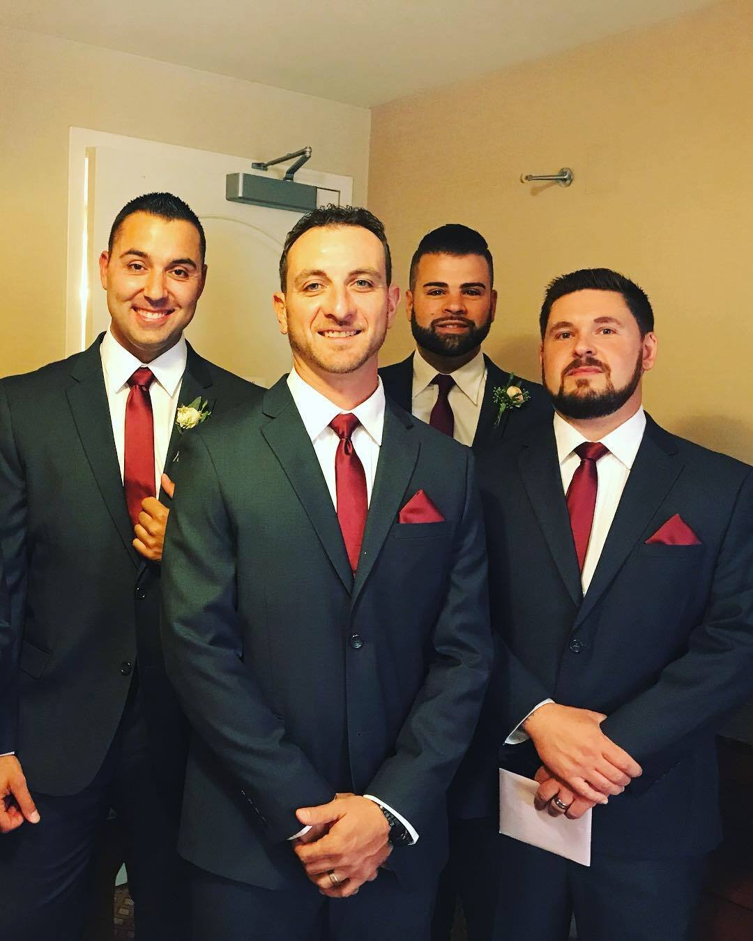 Four men wearing dark suits with red ties and pocket squares, standing in a room with a closed door.