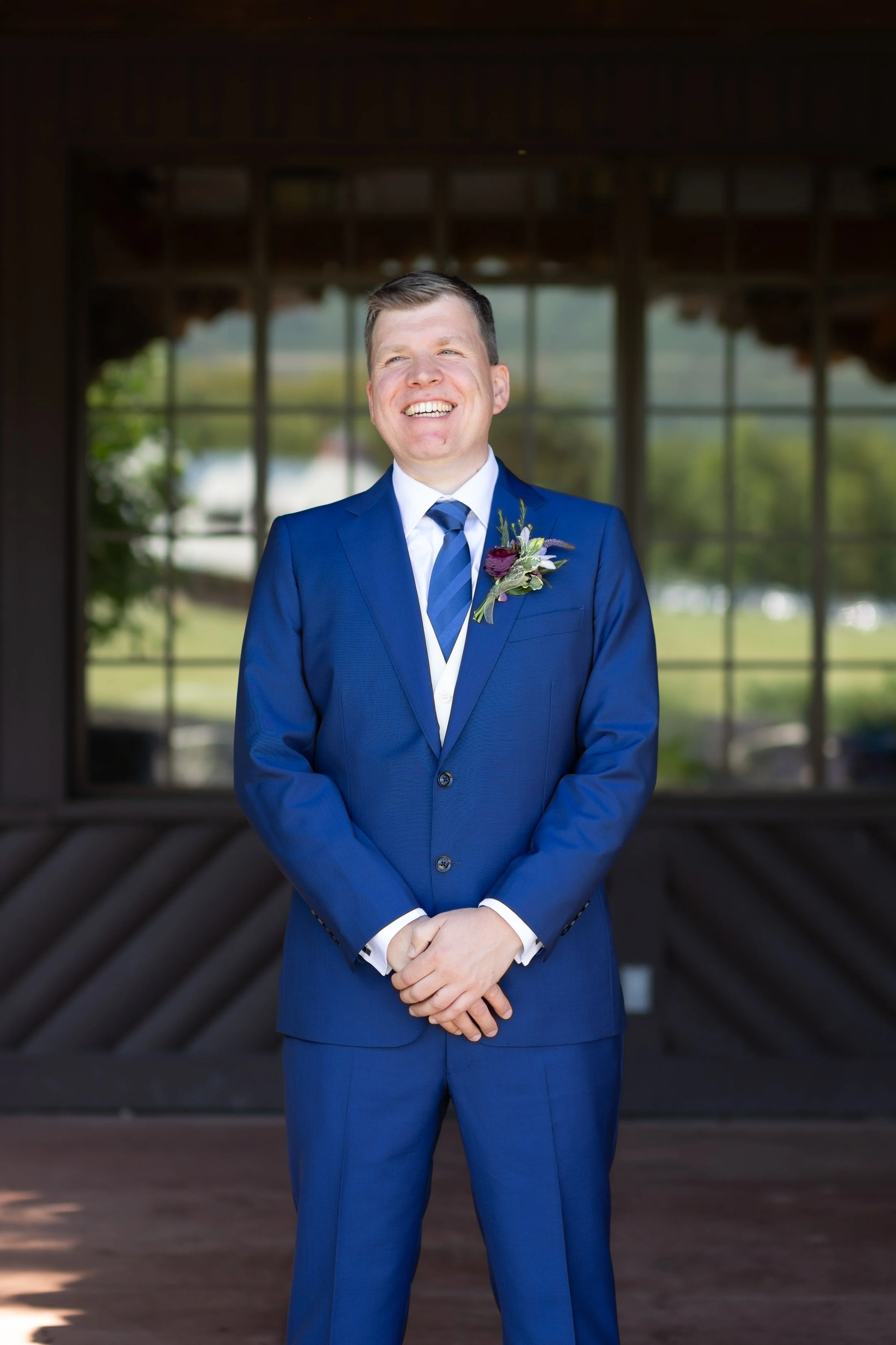 Smiling man in a blue suit with a boutonniere standing indoors at an event venue.