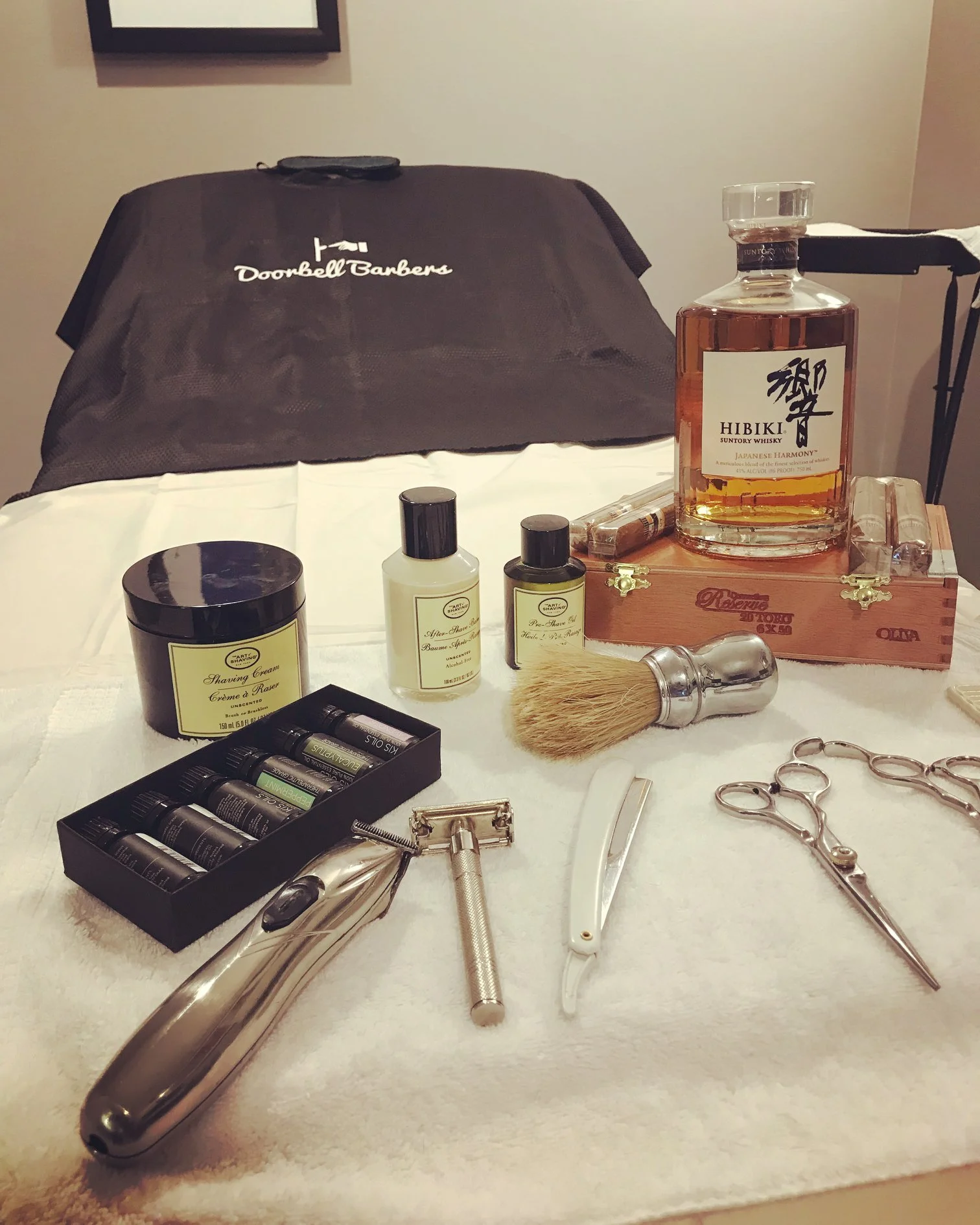 Barber tools and products on a table, including a clipper, straight razor, shaving brush, shaving cream, essential oils, and a bottle of Hibiki Japanese Harmony whiskey.