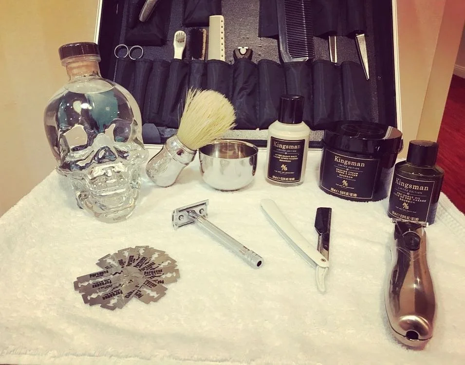 Shaving kit with items including a skull-shaped bottle, shaving brush, metal bowl, razor blades, safety razor, straight razor, electric trimmer, and Kingsman grooming products. Background features a set of grooming tools like scissors and combs.