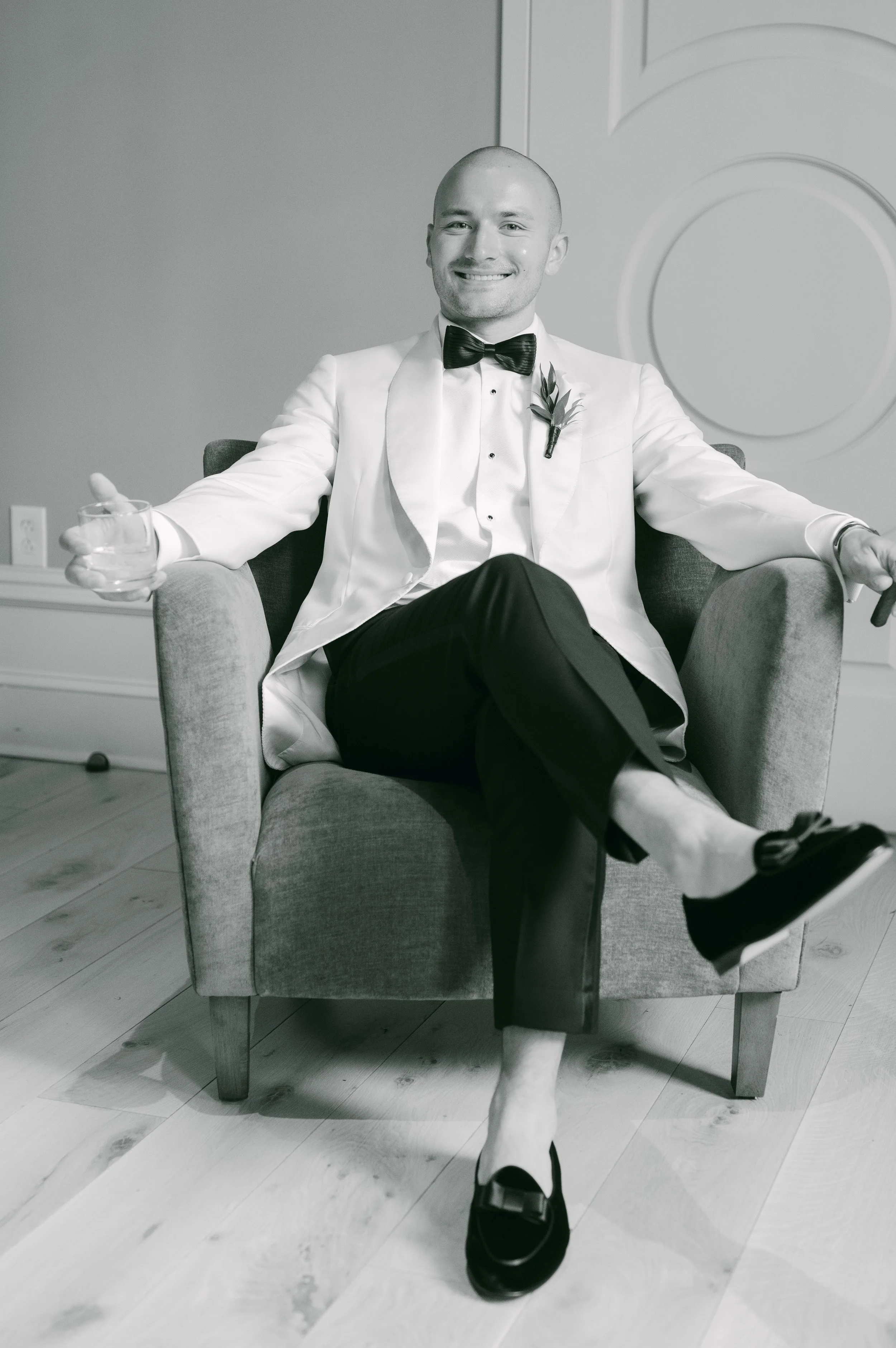 Black and white photo of a smiling man with a shaved head, dressed in a tuxedo with a bow tie and a boutonnière, sitting in a chair with one leg crossed over the other, holding a glass of drink in one hand, in a room with trimmed wood floors and pane