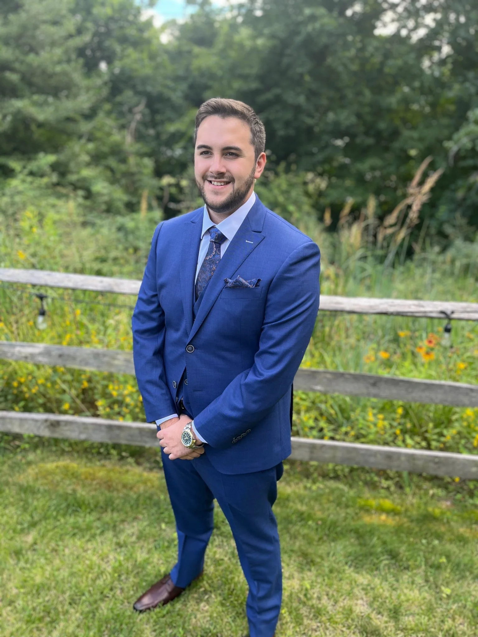 Person in a blue suit standing in front of a wooden fence with greenery in the background.