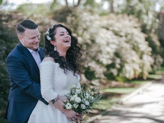 Bride and groom laughing outdoors in wedding attire; bride holding a bouquet.