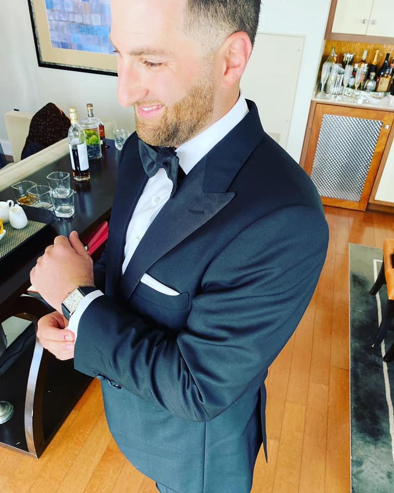 Man in a black tuxedo adjusting his cufflink, standing in a well-furnished room with bottles and glasses on a table.