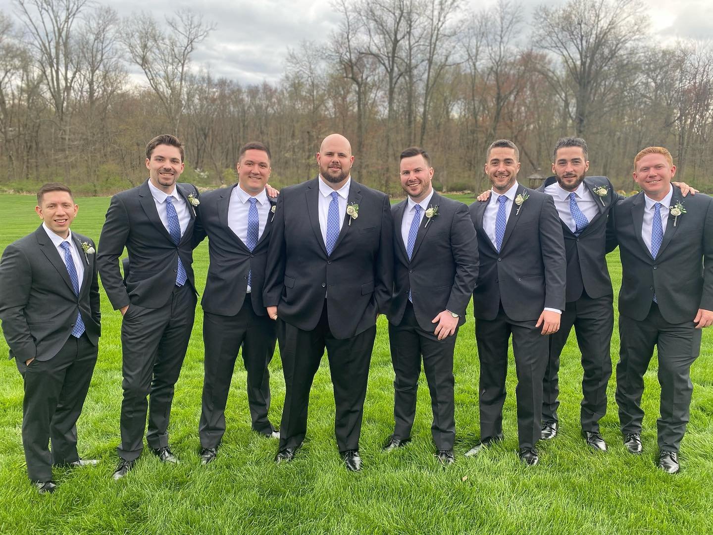 Group of seven men in matching black suits with blue ties and boutonnieres, standing on grass with trees in the background, overcast sky.