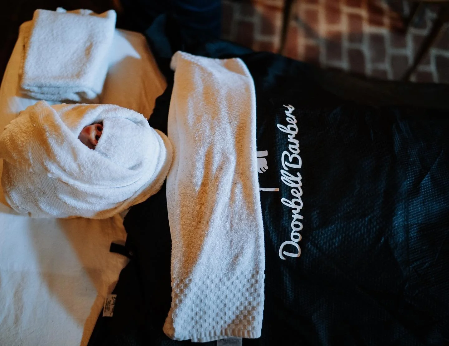 Person receiving a facial treatment with a towel wrap on the head and face, lying on a bed with "Doorbell Barbers" branding on a robe.