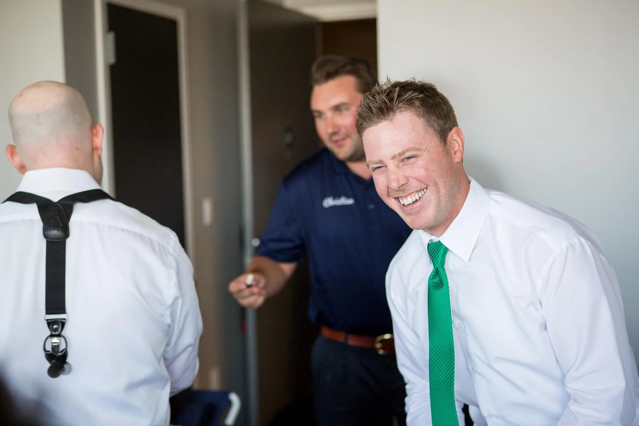 Three men getting ready together, one laughing, wearing white shirt and green tie.