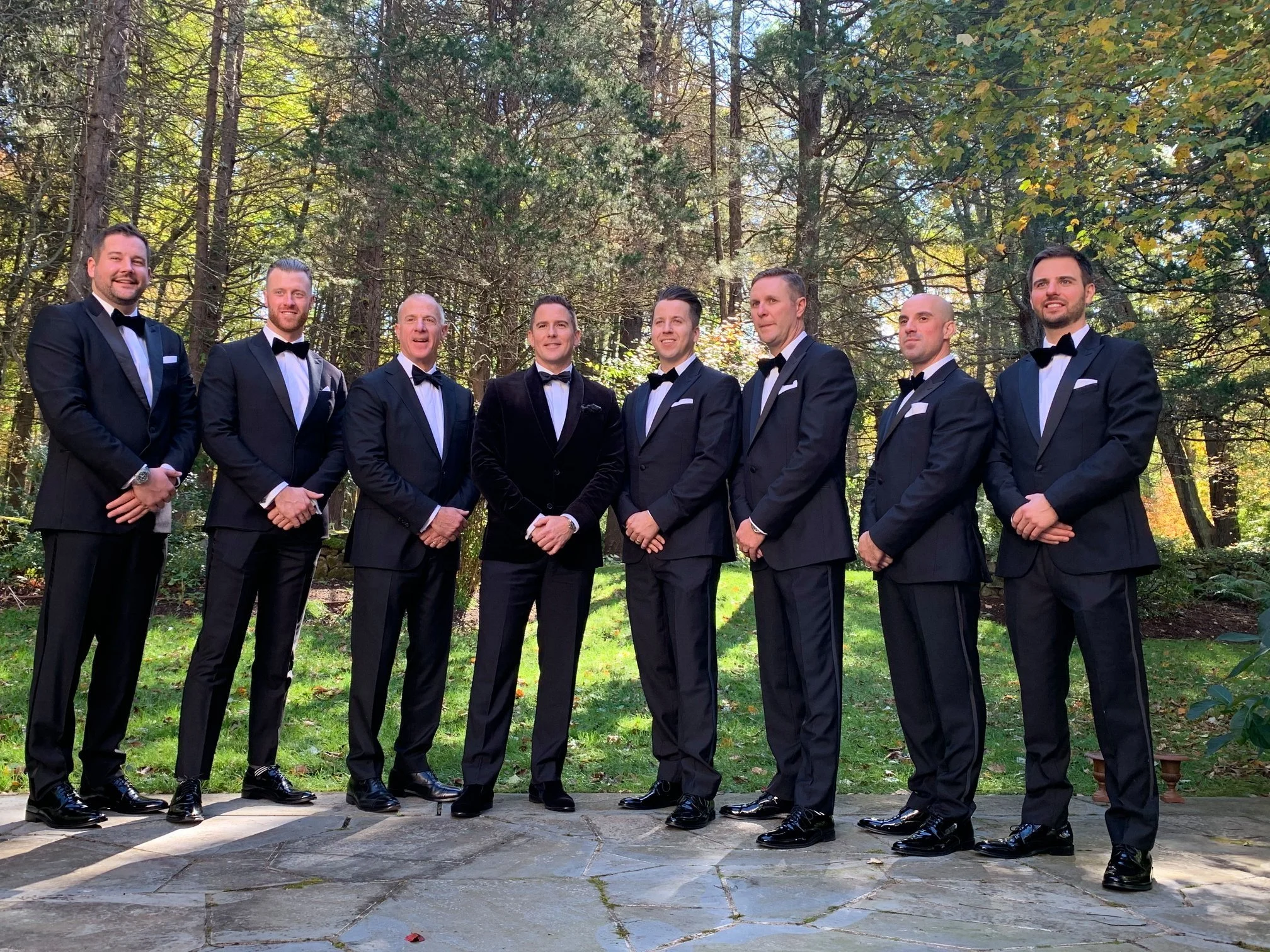 Group of men in tuxedos posing outdoors with trees in the background.