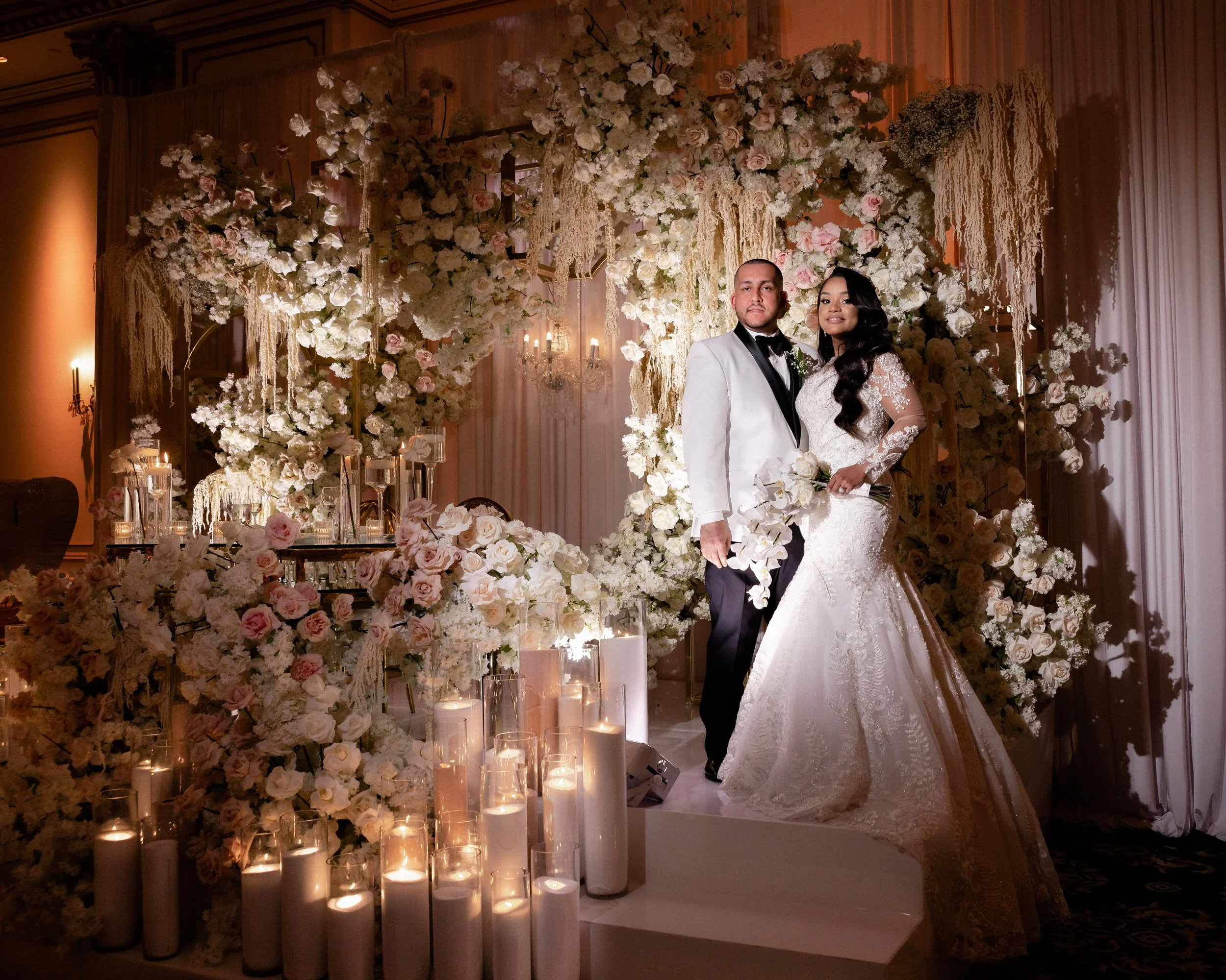 A bride and groom posing in an elegant wedding setup adorned with white and pink flowers, candles, and draped fabrics. The bride is in a detailed white gown, and the groom is in a white tuxedo jacket. The backdrop is lavishly decorated with floral ar