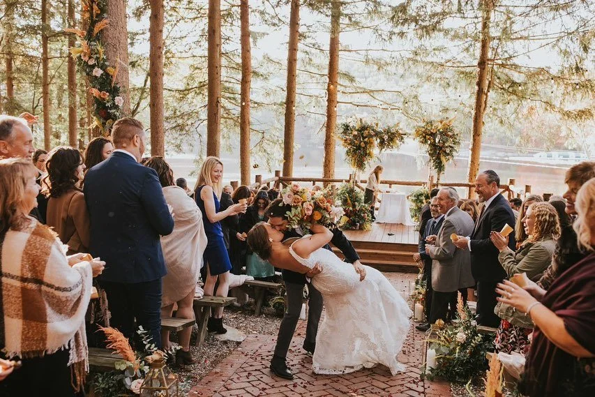 Newlywed couple kissing at an outdoor wedding ceremony surrounded by guests in a forest setting.