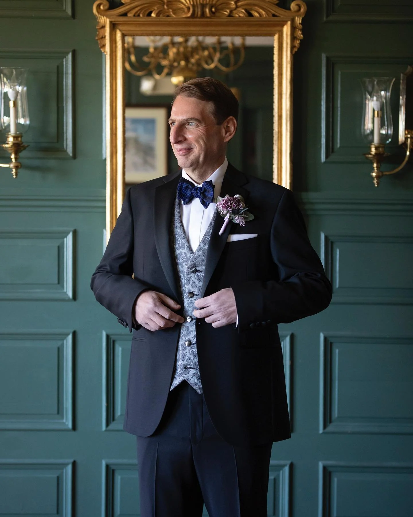 Man in formal tuxedo with a blue bow tie and boutonniere standing in front of a green paneled wall with a gold-framed mirror.