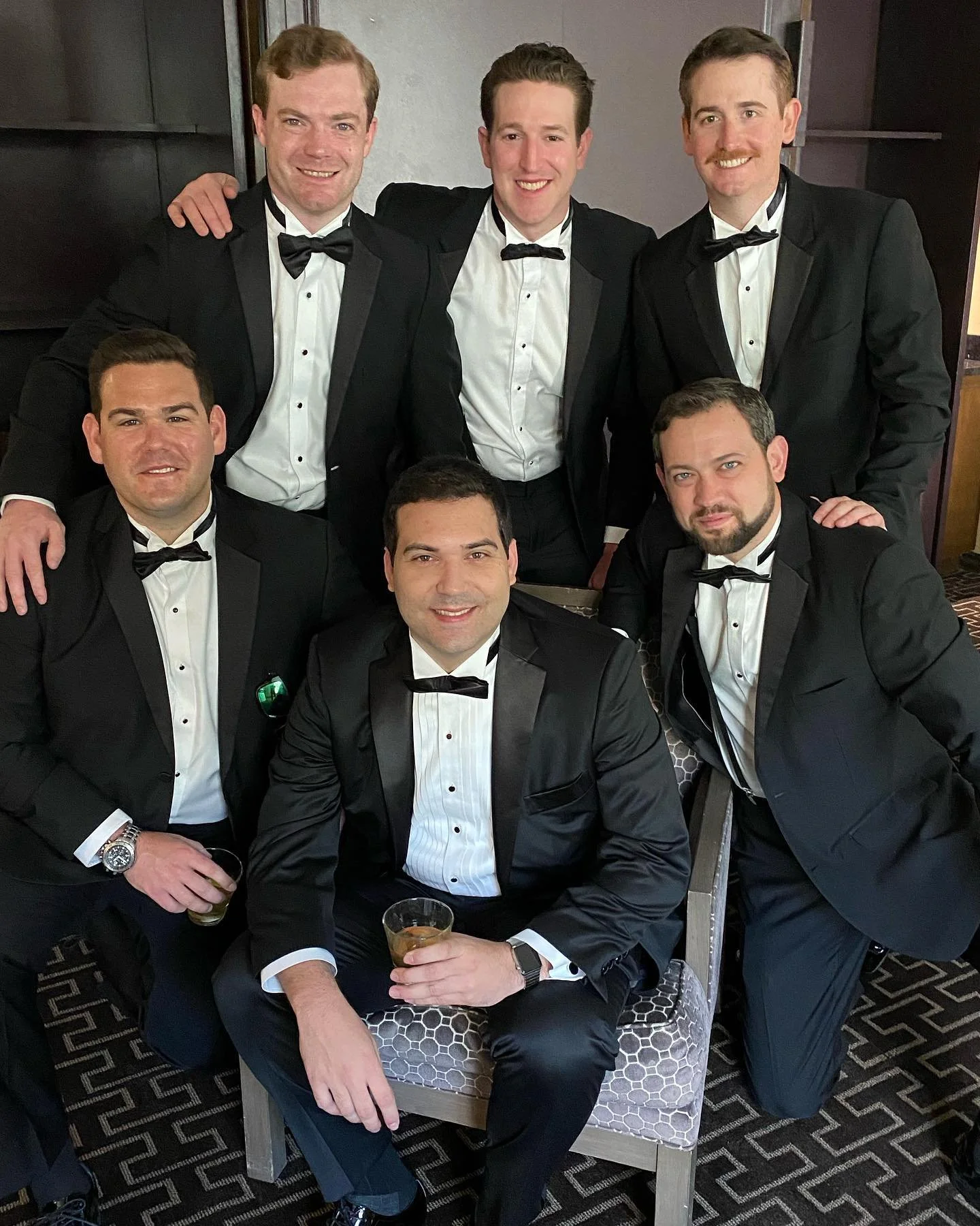 Group of six men wearing black tuxedos with bow ties, posing indoors.