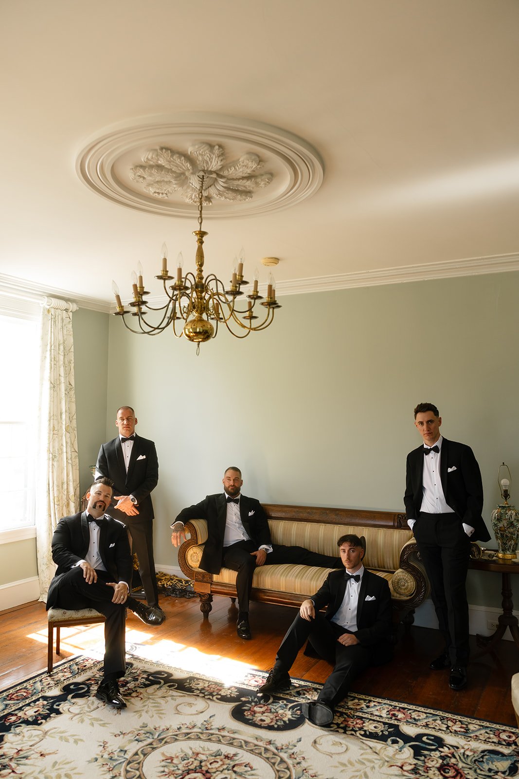 Five men in tuxedos in a decorated formal room, some sitting and some standing, with a chandelier and vintage furniture.