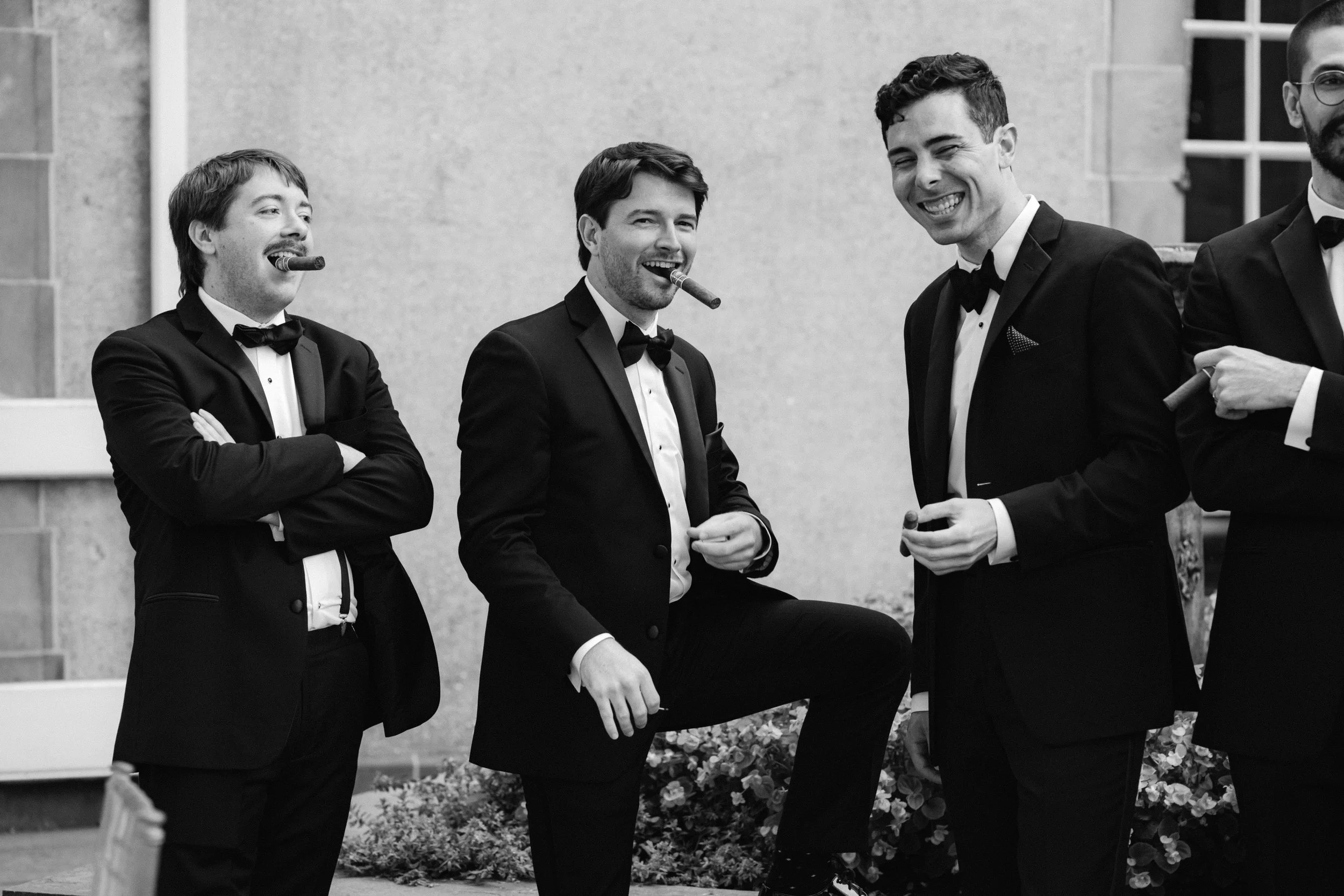 Group of men in tuxedos with cigars, laughing outdoors, black and white image.