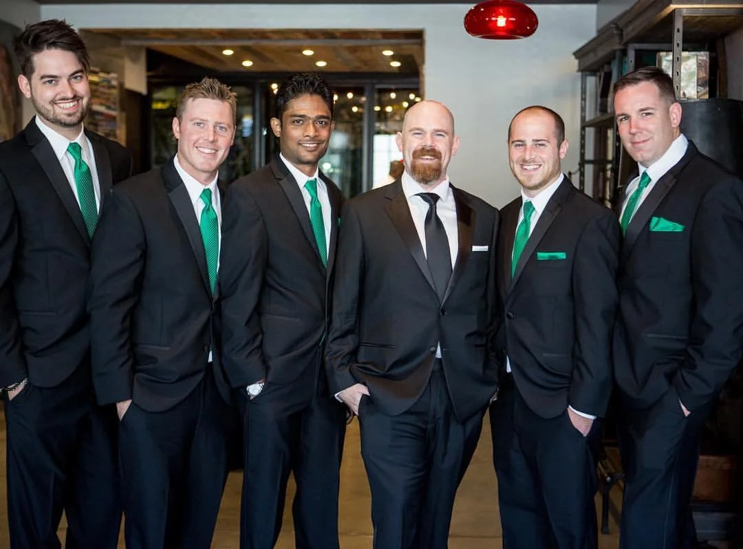 Group of six men in formal black suits with green ties and pocket squares, posing indoors with smiles.