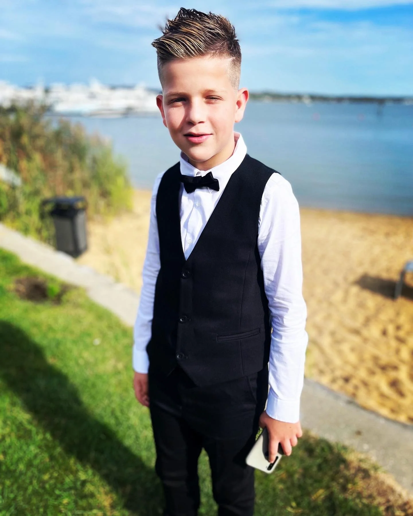 Boy wearing formal attire with a bow tie standing by a waterfront on a sunny day.