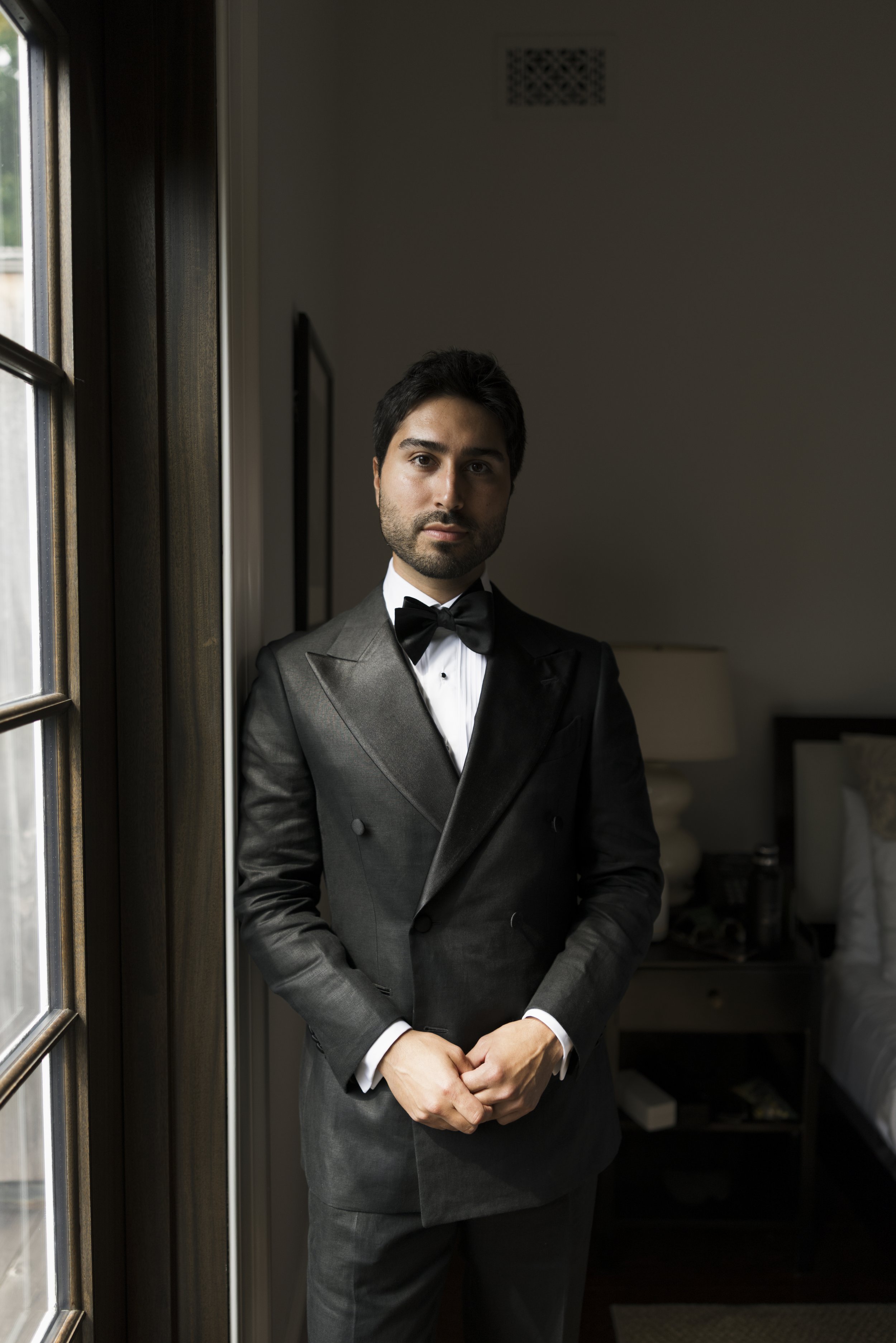 A man in a black tuxedo with a bow tie standing in a softly lit room, near a window, with a serious expression.