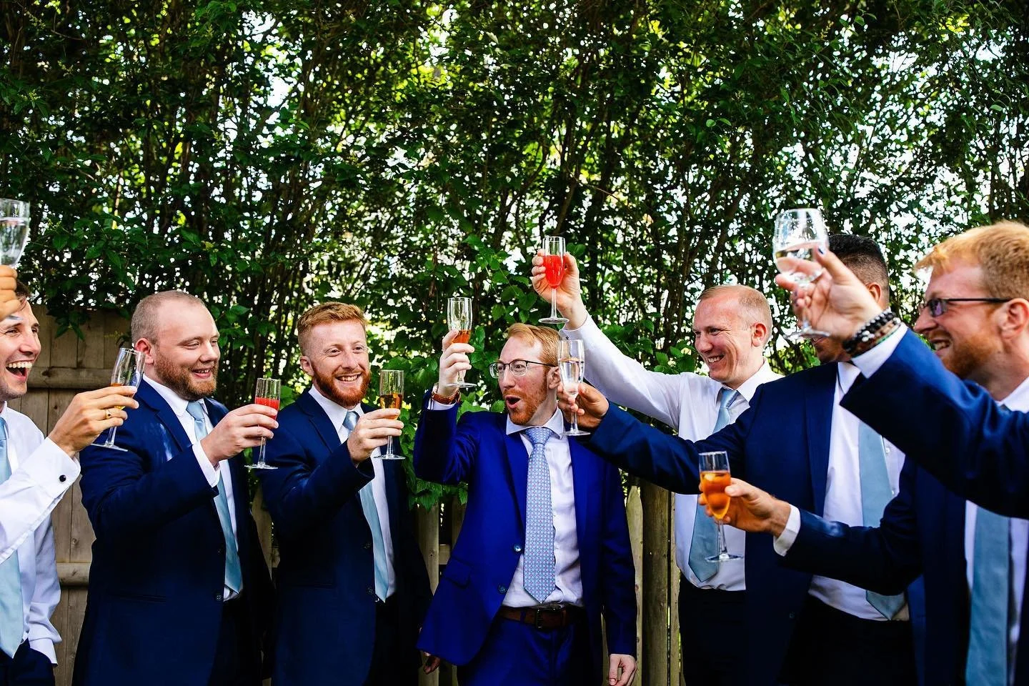 Group of men in suits holding up glasses in a celebratory toast outside with greenery in the background.