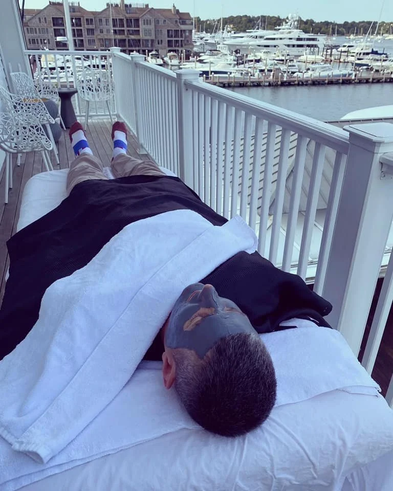 Person lying on a massage table with a face mask, outdoors on a balcony overlooking a marina with boats.